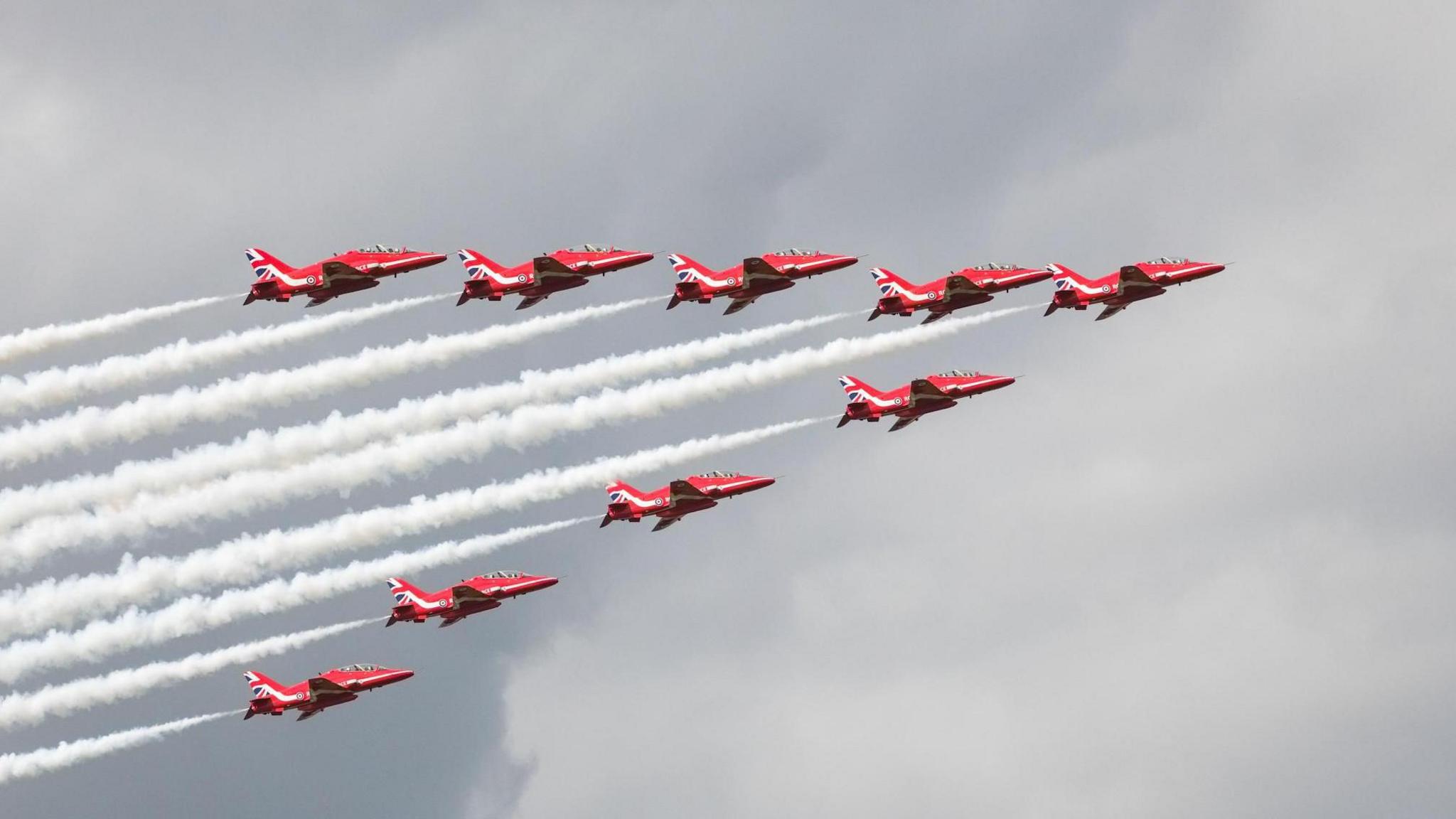Nine Red Arrow jets in a triangular formation, leaving smoke trails in their wake. The red planes have Union Jack livery on their tails.