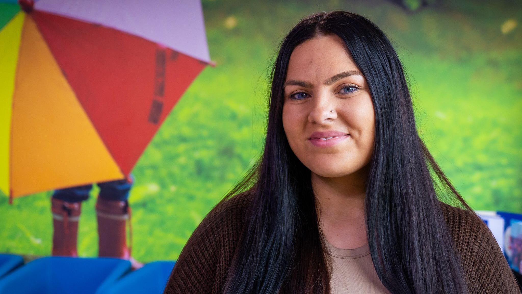 A girl with long brown hair, smiling at the camera. Behind her is a picture of a person under a multi-coloured umbrella standing on the grass, wearing wellies.