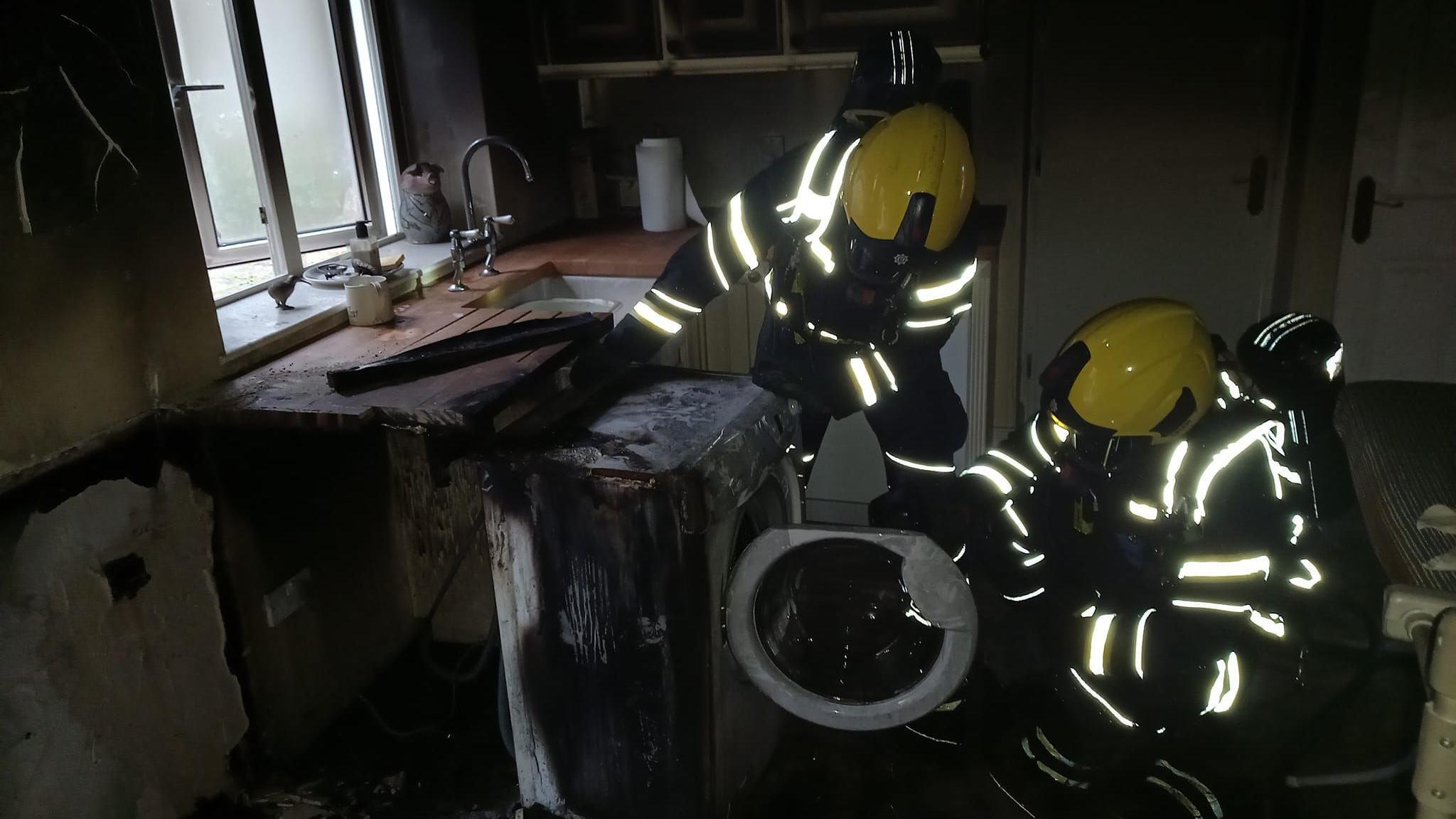 Two firefighters in protective clothing and wearing yellow helmets inspect a burnt tumble dryer following a blaze which has caused significant fire damage to the room it is in.