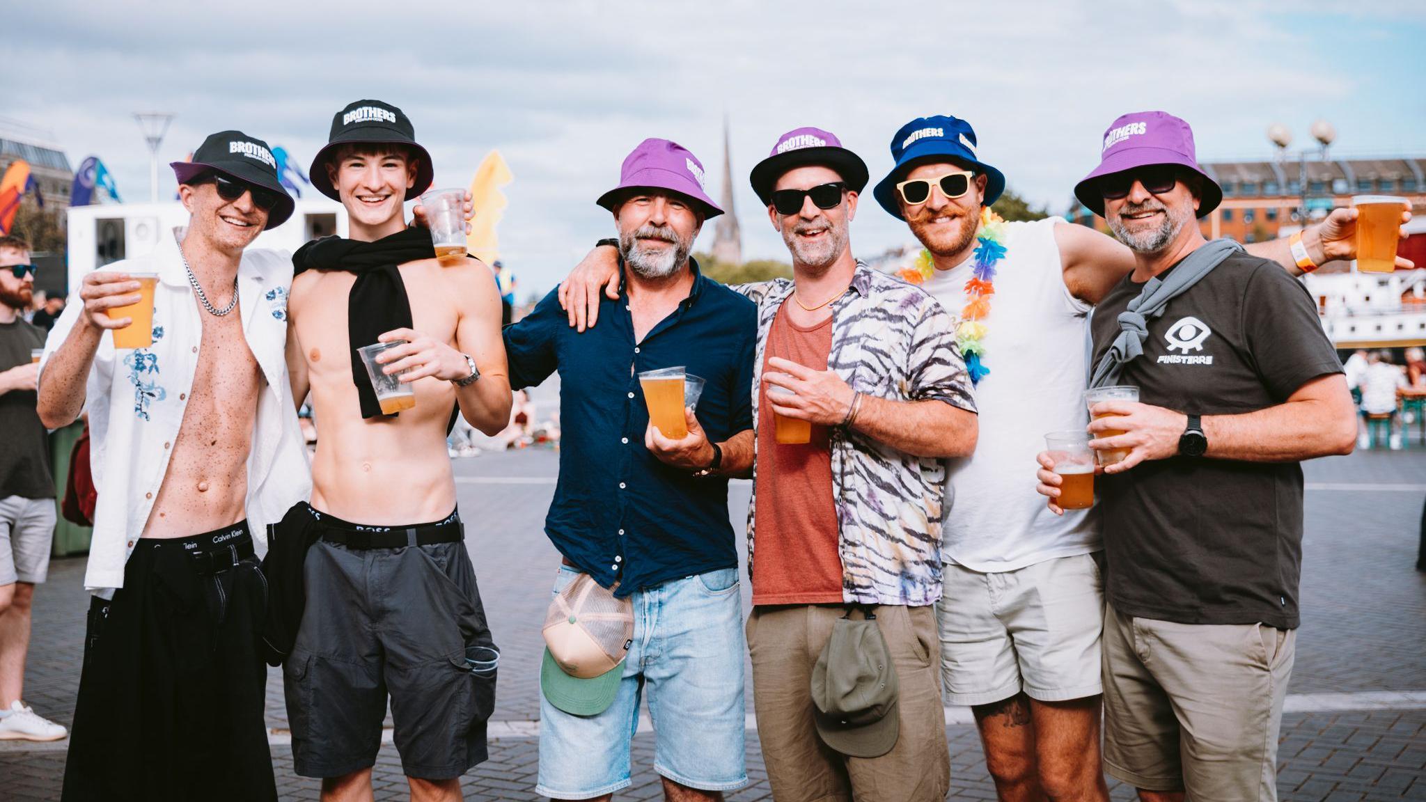 A group of men in a huddle looking at the camera. They are all wearing bucket hats and holding plastic cups full of beer. They are all smiling at the camera