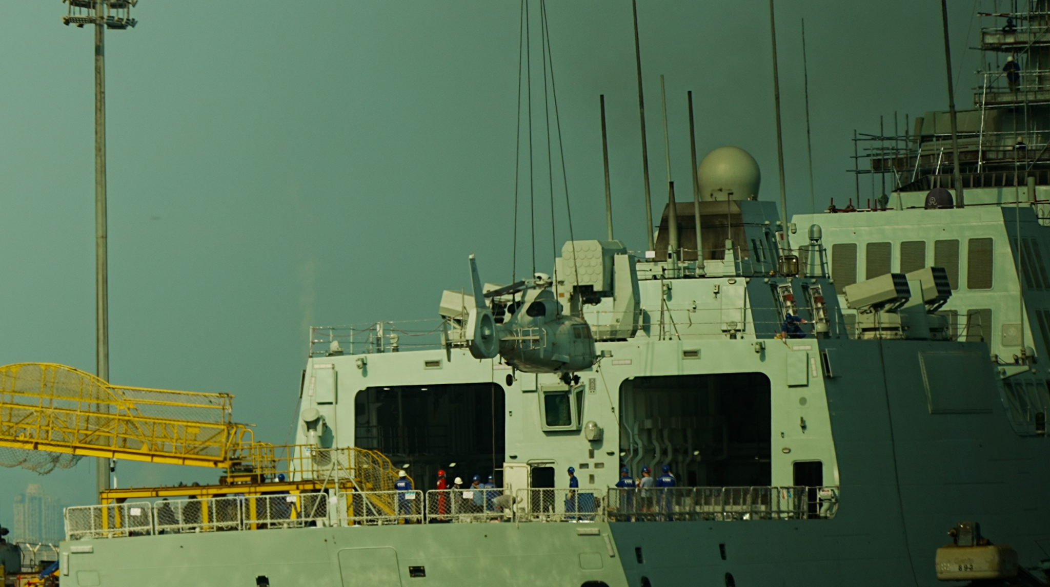 A helicopter is being lowered onto the deck of a military vessel by a crane, while the crew watch.