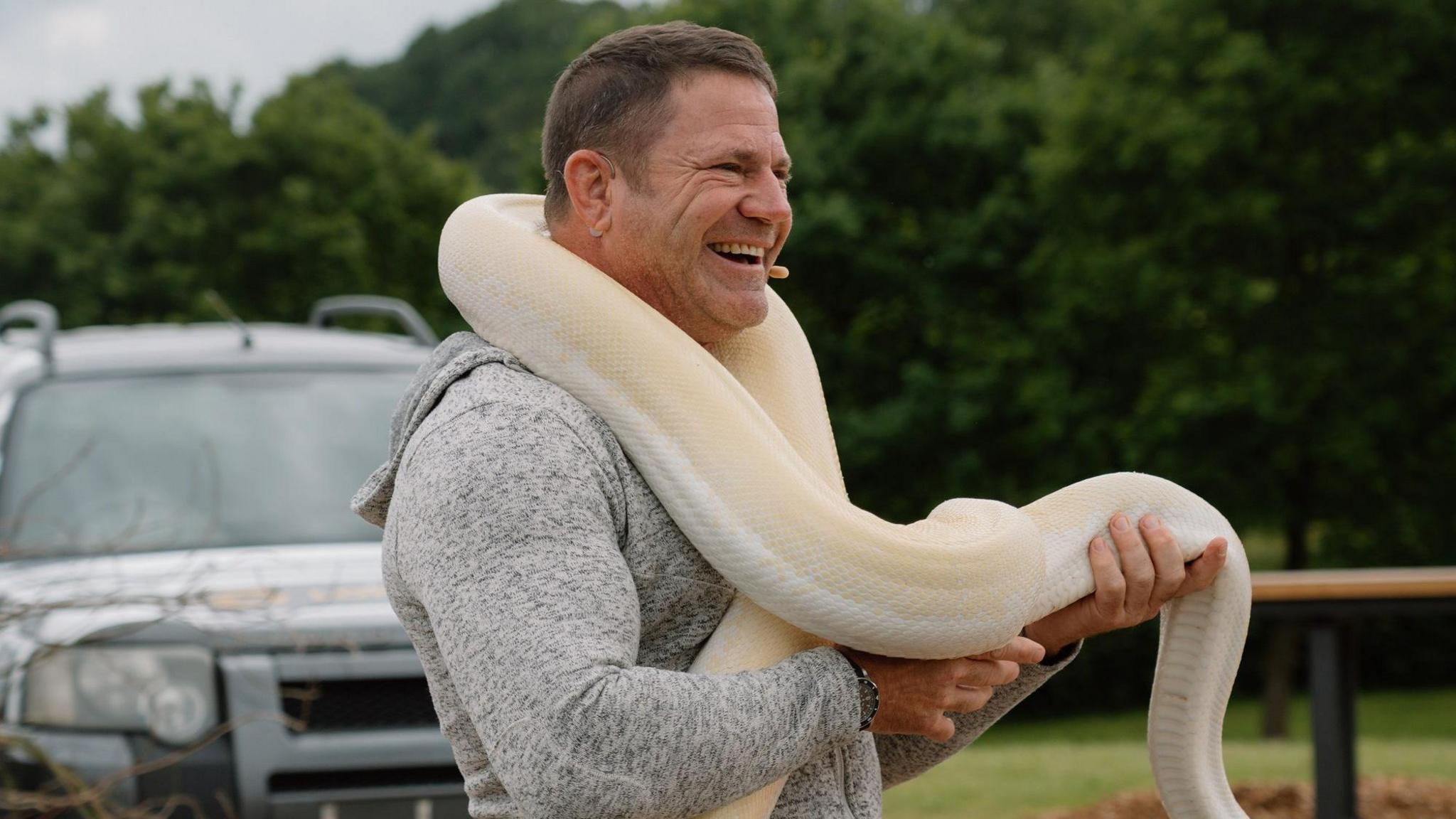 A side shot of Steve Backshall laughing in a grey hoodie with a massive cream-coloured snake around his shoulders and arms.