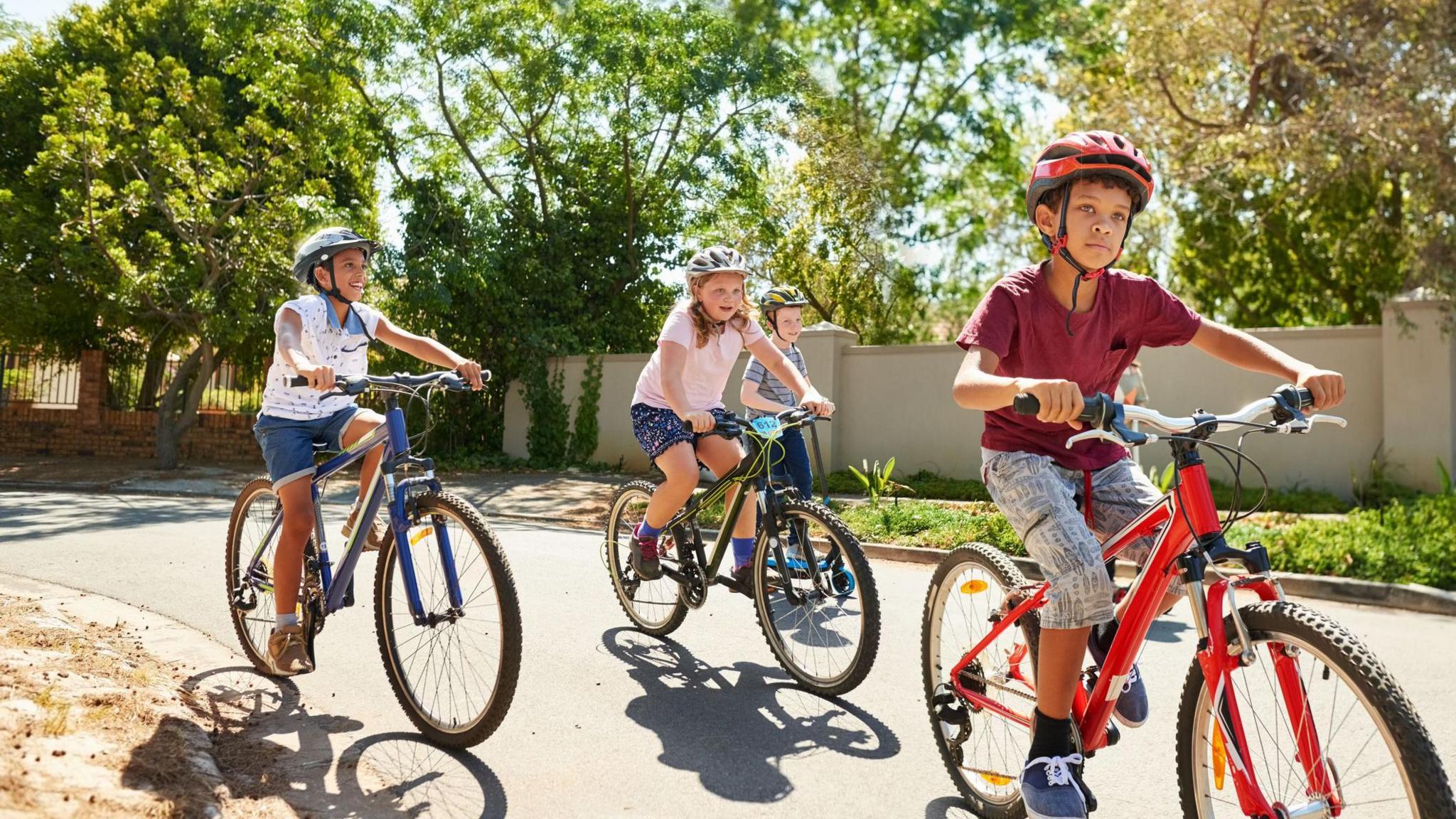 Children on bikes cycle along the road, it's sunny and they are all wearing helmets