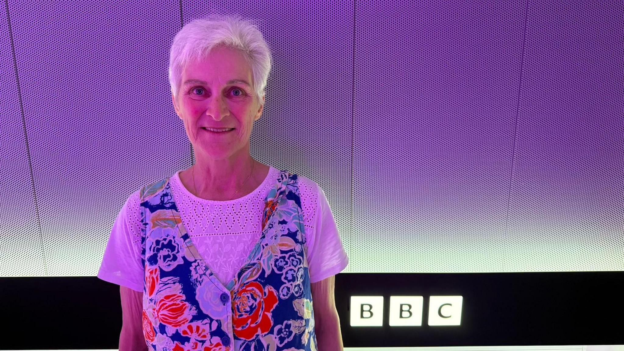 Fiona is standing in front of a purple background in a BBC building. She has short white hair and is wearing a white T-shirt under a blue, white and red tunic dress.