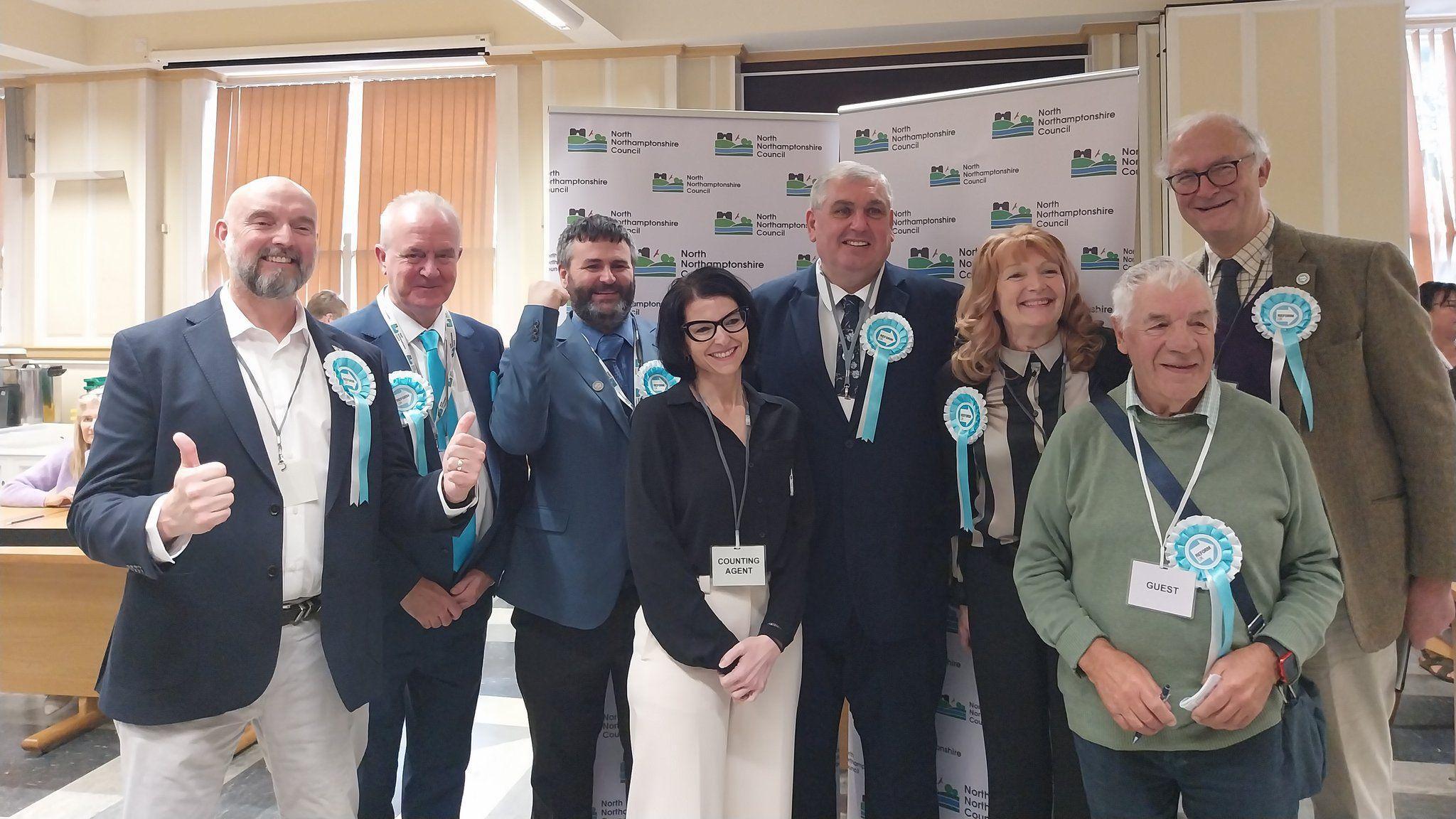 Eight Reform UK supporters, all looking to the left, mostly wearing turquoise rosettes. They are standing by a council pull-up sign, and are all smiling.