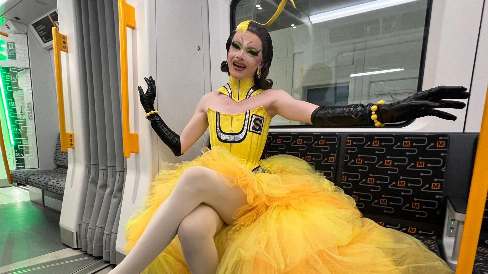 Sally TM sitting on a Metro seat with her arms outstretched. She is smiling into the camera with her mouth open. She is sitting on the dark Metro seats with the yellow logo.