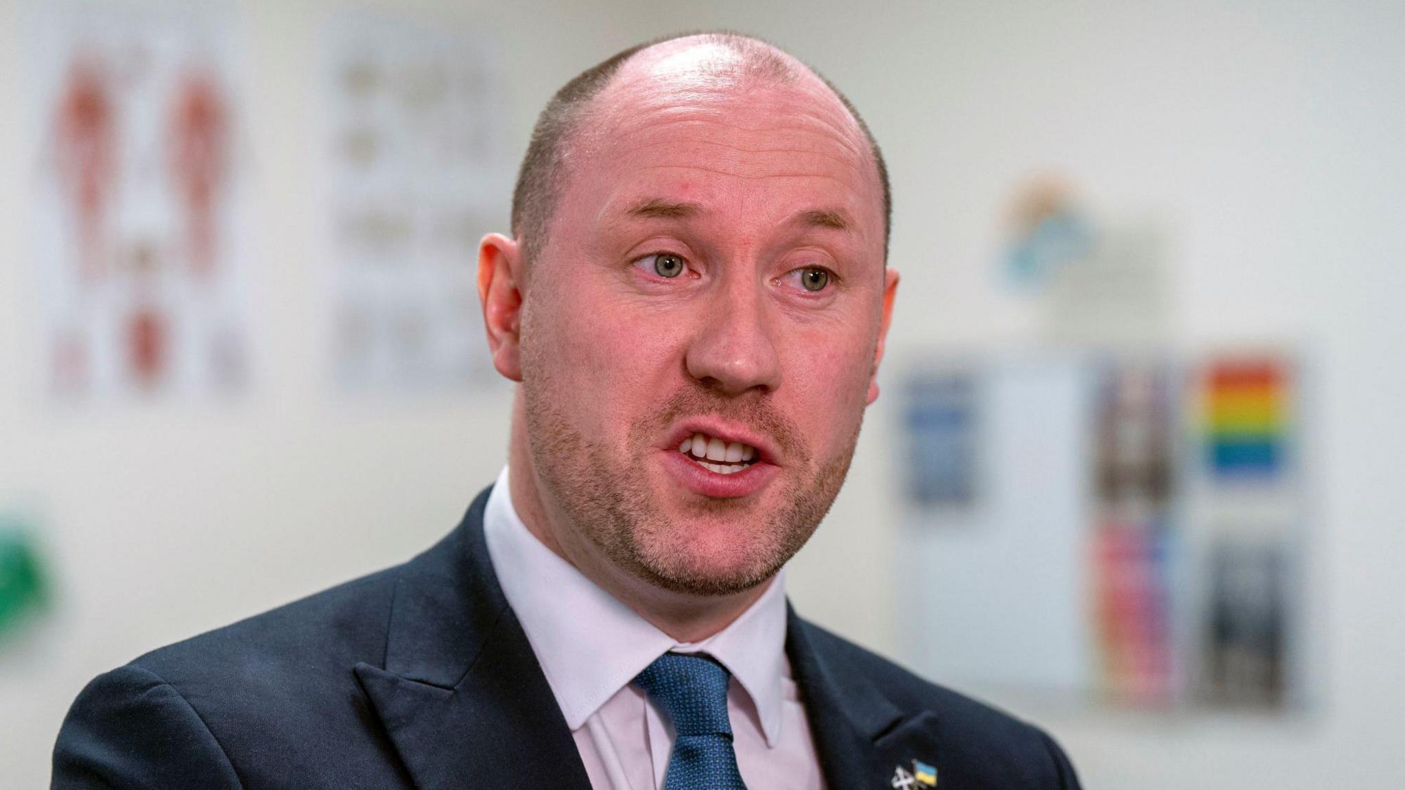 Health Secretary Neil Gray, wearing a dark suit, pink shirt and navy tie, is pictured in a medical setting - posters on the walls behind him blurred.