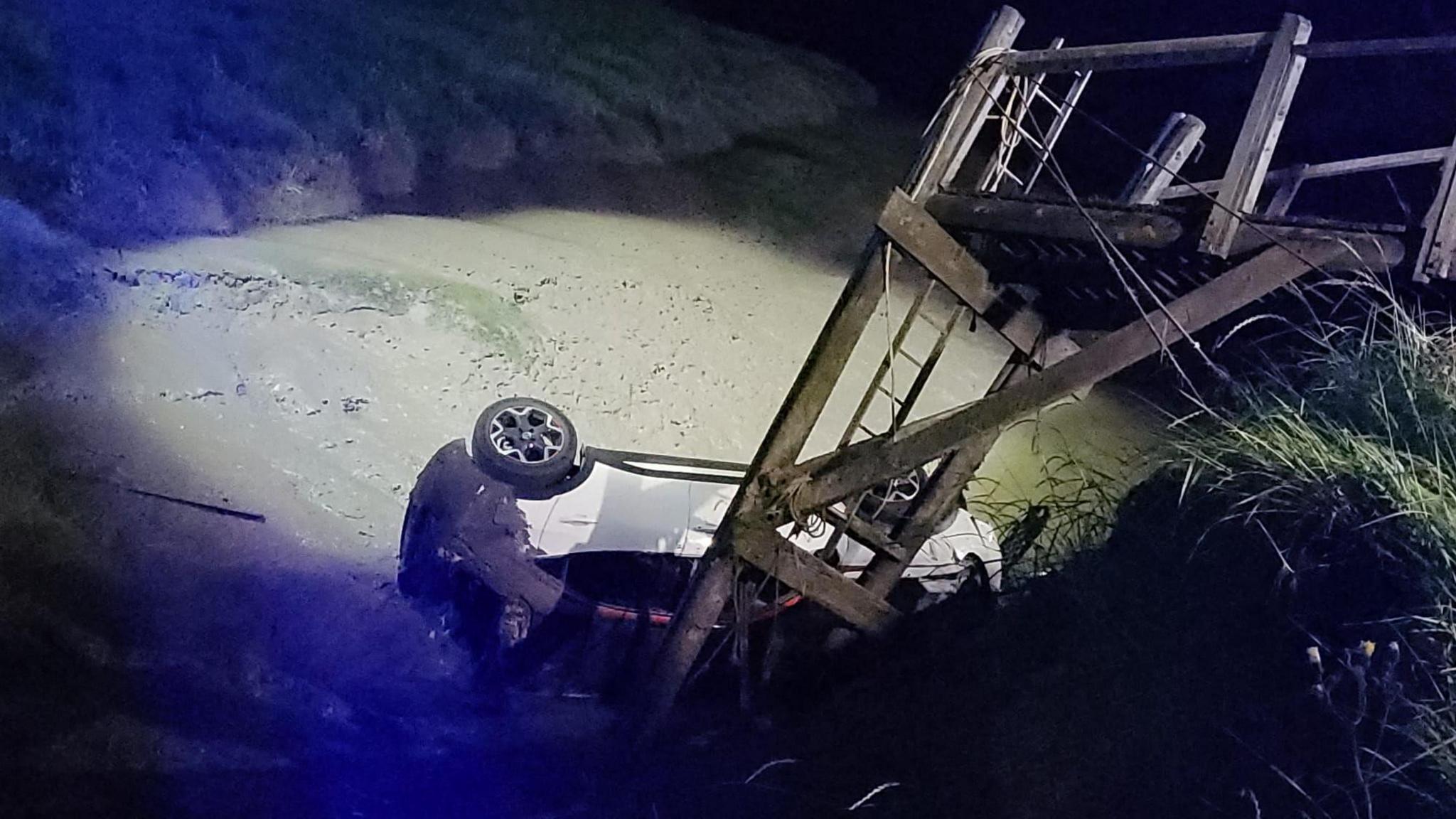 A car is on its side in a creek, surrounded by mud, at nighttime