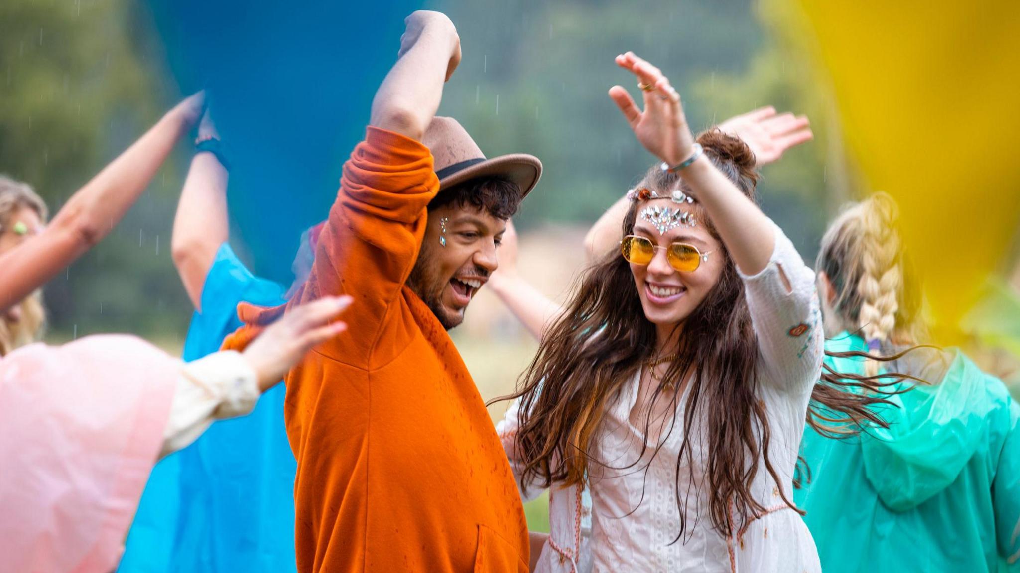 A man and a woman at a festival wearing glitter and gems on their faces and dancing while it rains slightly