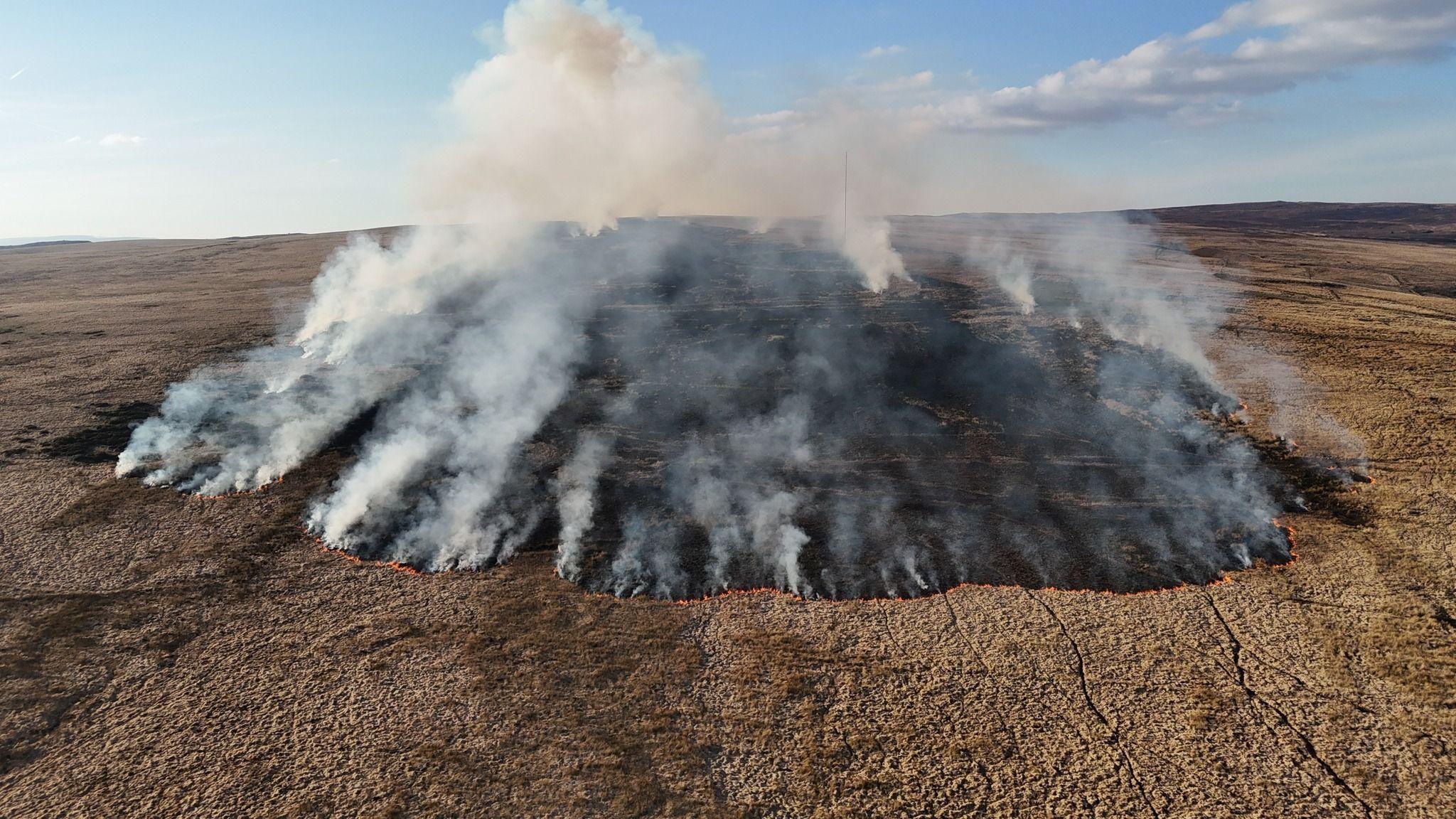 An aerial picture of a large mountain fire actively burning in a large circular shape. Lots of grey smoke is rising into the sky, leaving a blackened patch on the ground.