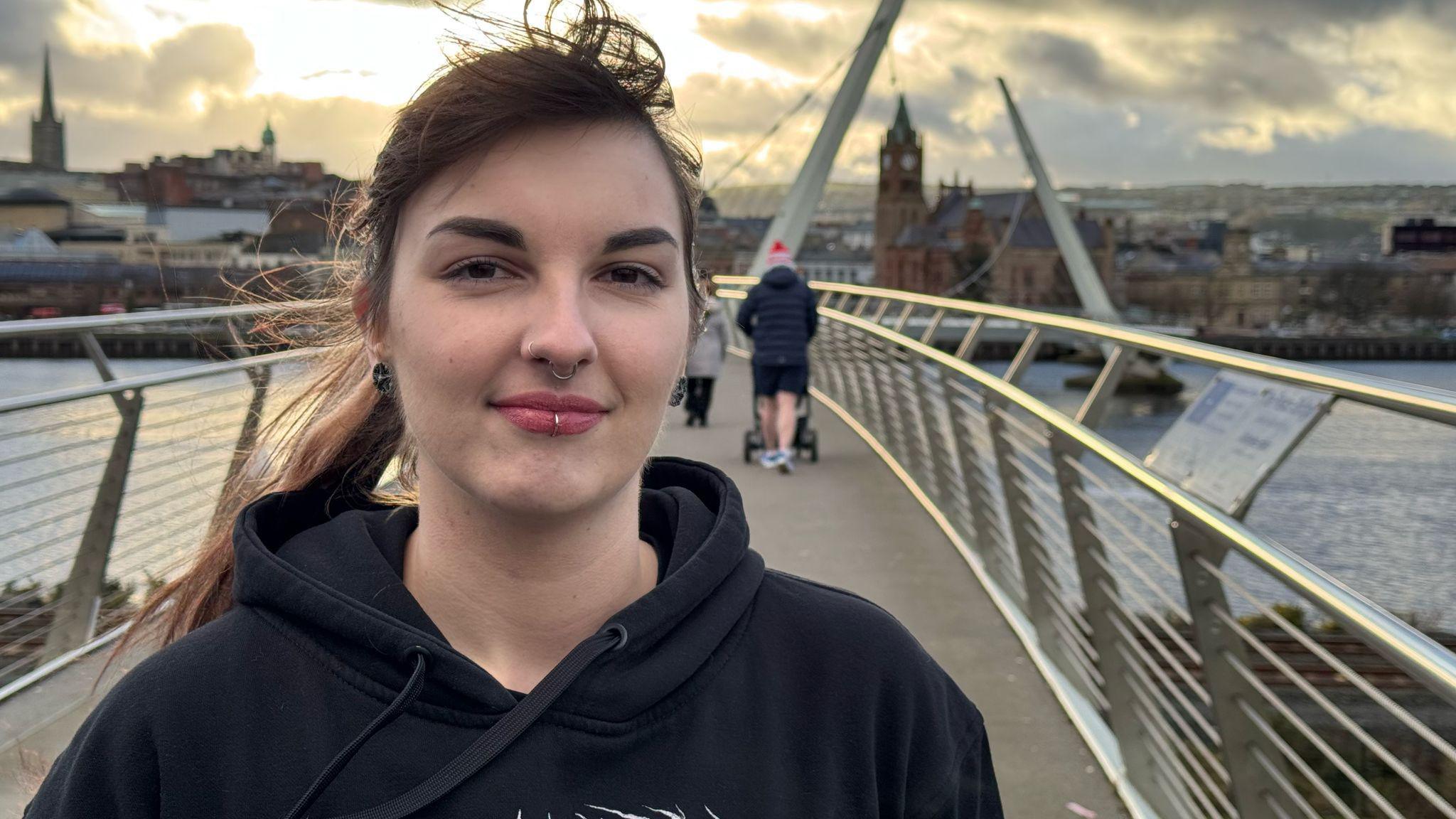 Alannagh Doherty, a woman with long, brown hair, a lip piercing and two nose piercings. She is standing on a silver railed bridge wearing a black hoodie. Behind her is Derry city.  A man in a blue coat and shorts is walking away from her, pushing a buggy, he has on a red hat.  