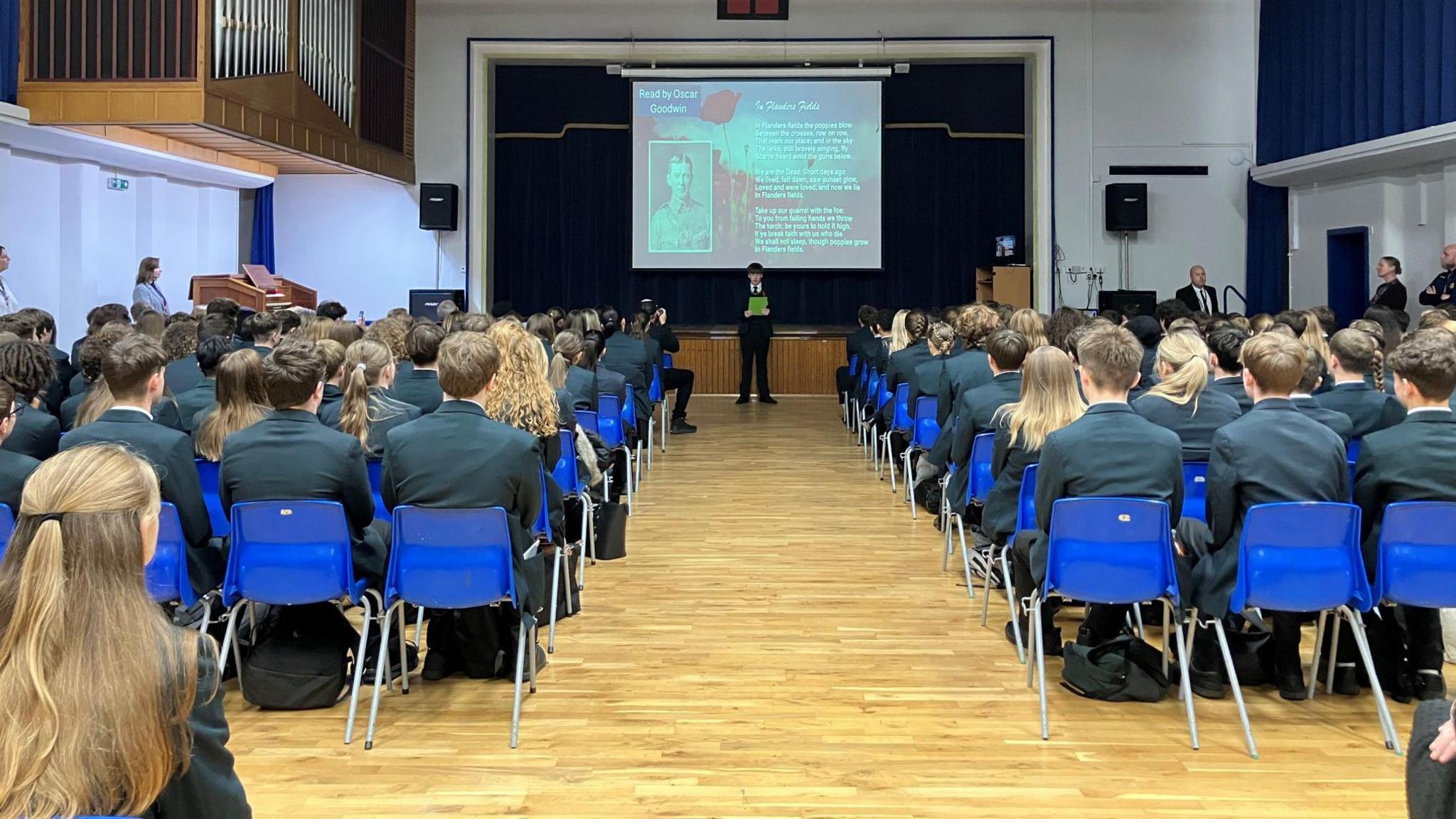 Rows of bright blue plastic chairs  separated by a central aisle. The chairs are mostly occupied by students wearing dark blazers, all facing forward toward a stage area. At the front, a male student is stood holding a sheet of green paper. Behind him there is a large projection screen with the image of a man and words on.