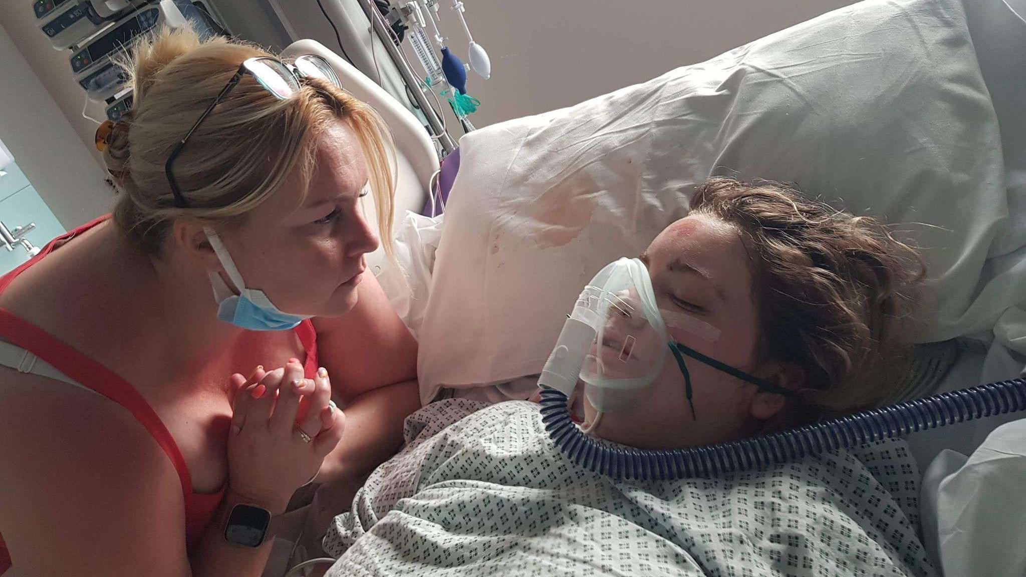 A woman in a hospital bed has her eyes closed and is wearing an oxygen mask. She has brown hair and has a cut on her forehead. A woman is sat next to her with her hands together in a praying motion. She has blonde hair and is wearing a red top.