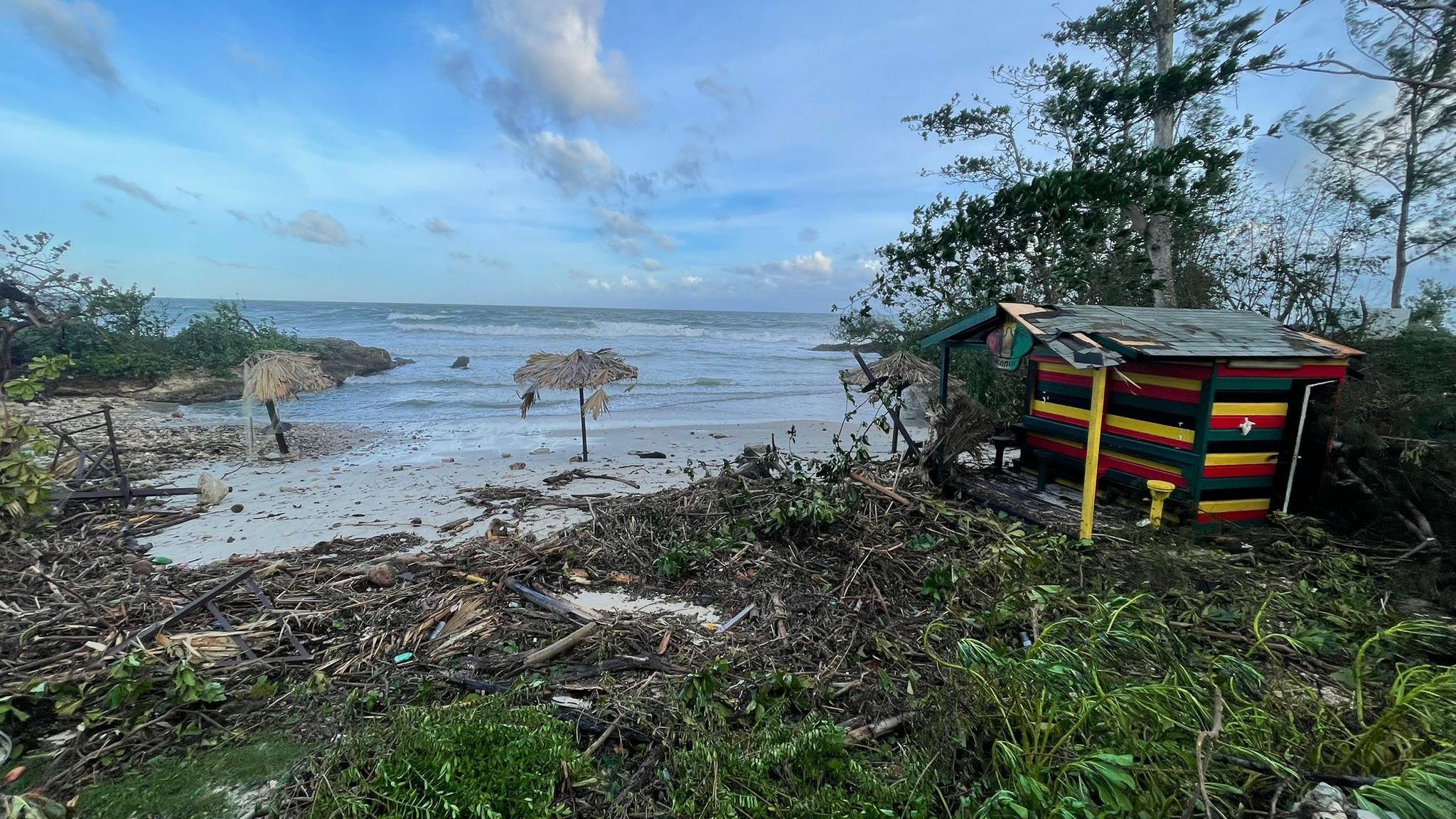 A white sand beach littered with debris, and umbrellas partially destroyed, next to a small hut painted in the Jamaican flag colours. A blue sky has scattered grey clouds and grass and trees surround the beach.