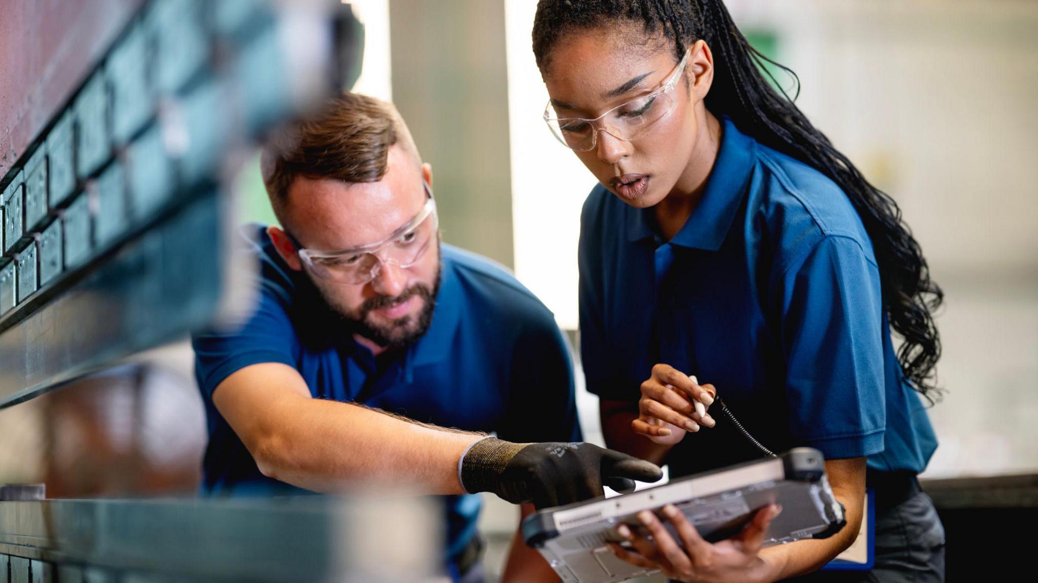 A young woman and a man look at a tablet in a car factory. They are wearing blue polo shirts and safety goggles. He is wearing black gloves and pointing at the tablet.