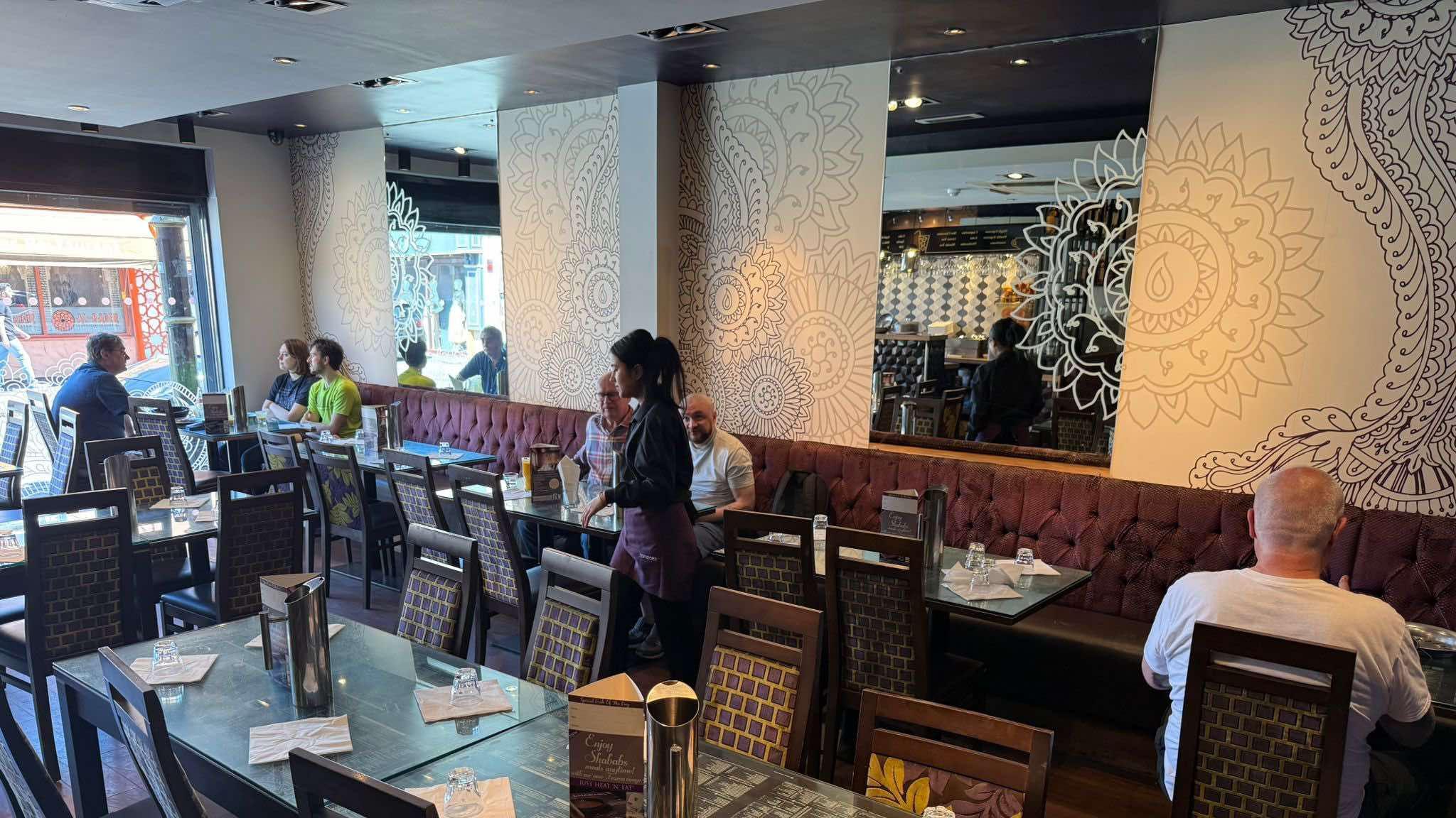 An Indian restaurant with customers sat waiting to eat their meals. A female employee in a purple uniform can be seen walking around. There are patterns on the wall and mirrors.