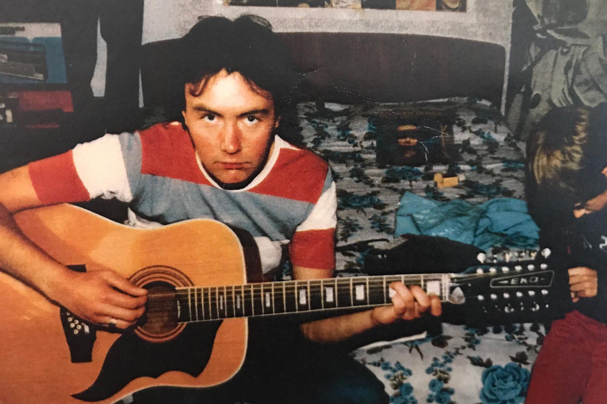 Irvine Welsh looking to camera in red, white & check T shirt playing the guitar