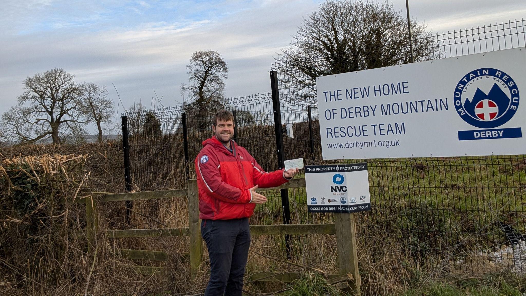 A man with a red jacket holding up a cheque in front of a sign which reads "the new home of Derby Mountain Rescue Team".