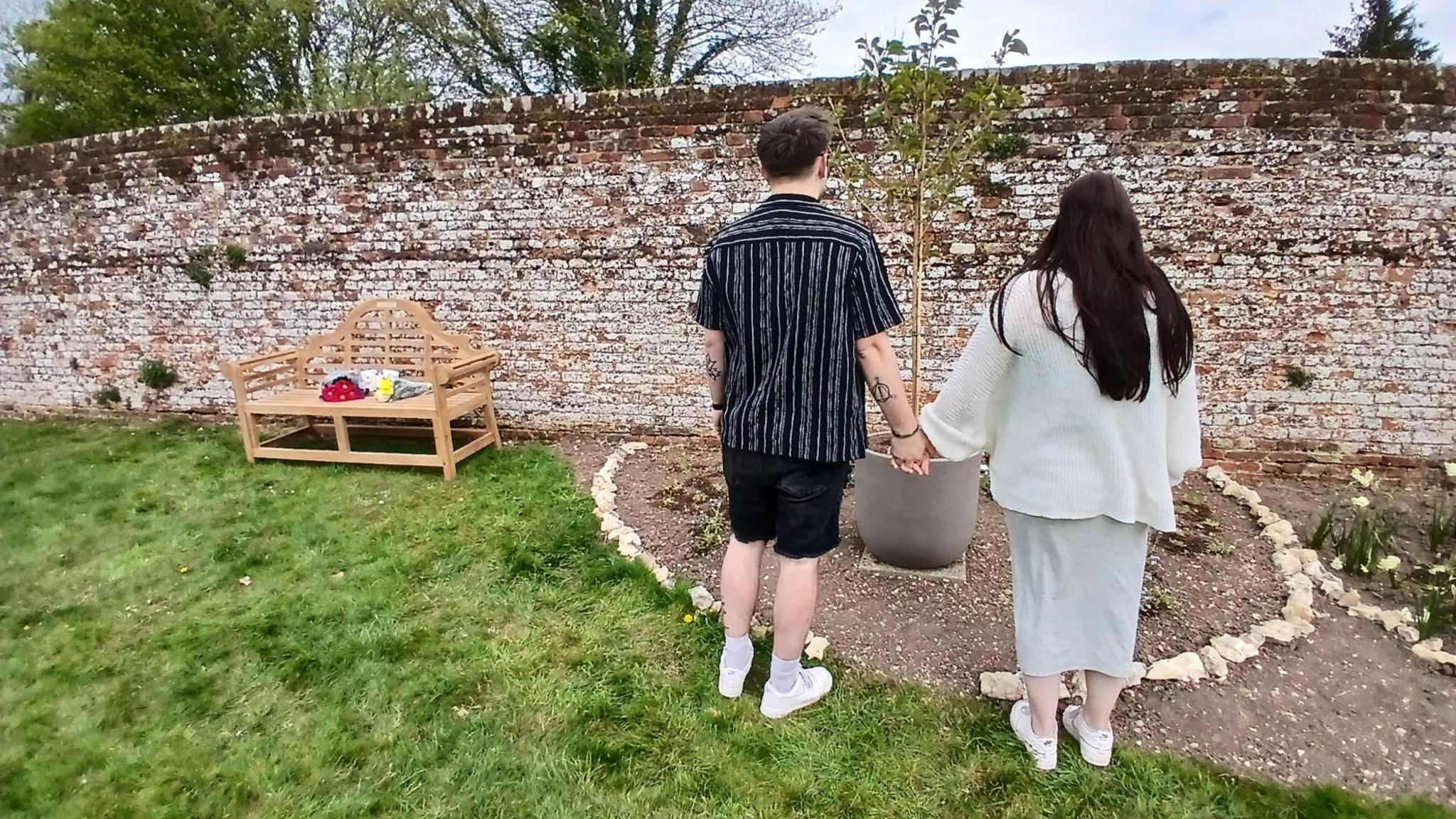 A man and a woman stood next to each other holding hands and looking down at a small tree in a pot which is flanked by white rocks in a green garden. There is a bench with bunches of flowers next to them.