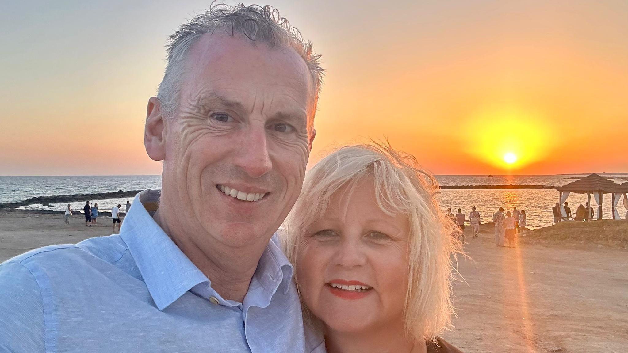 Stuart Emerson standing on the beach beside his wife Claudia. He has short white hair and is wearing a light-blue collared shirt, smiling at the camera. Claudia has shoulder-length blonde hair and is wearing red lipstick, also smiling at the camera. Behind them the sun is setting on the horizon and the whole sky is lit up orange.