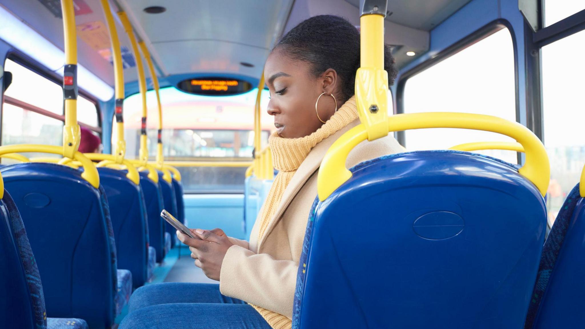 A young woman with her hair tied back sitting sideways on a bus looking down at her smartphone