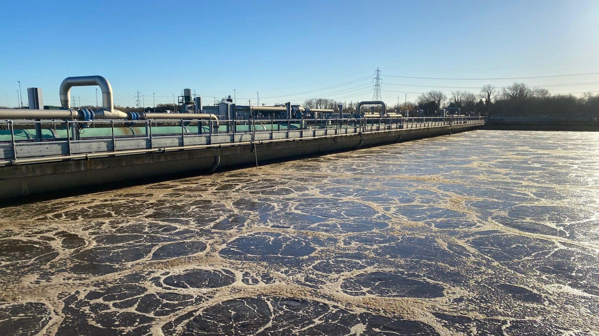 Sewage being treated at a Severn Trent waste water plant. Light brown circles of foam can be seen on top of brown water which fills most of the frame. Along one side of the water is a concrete bank with a series of pipes running along the top. A pylon carrying electricity cables is visible in the background.