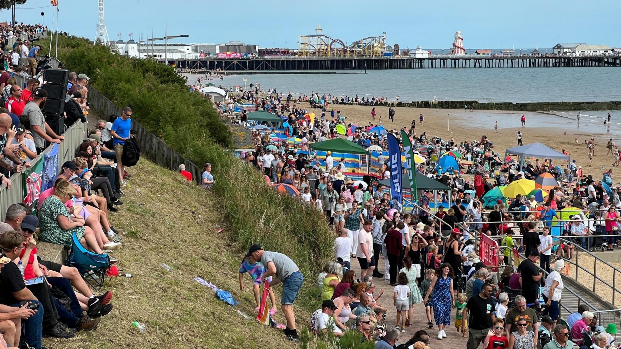 Crows of people line a seafront, sitting on grass banks and standing along the beach and promenade area to wait for aerial displays. A pier is in the distance with a rollercoaster and a helter skelter.