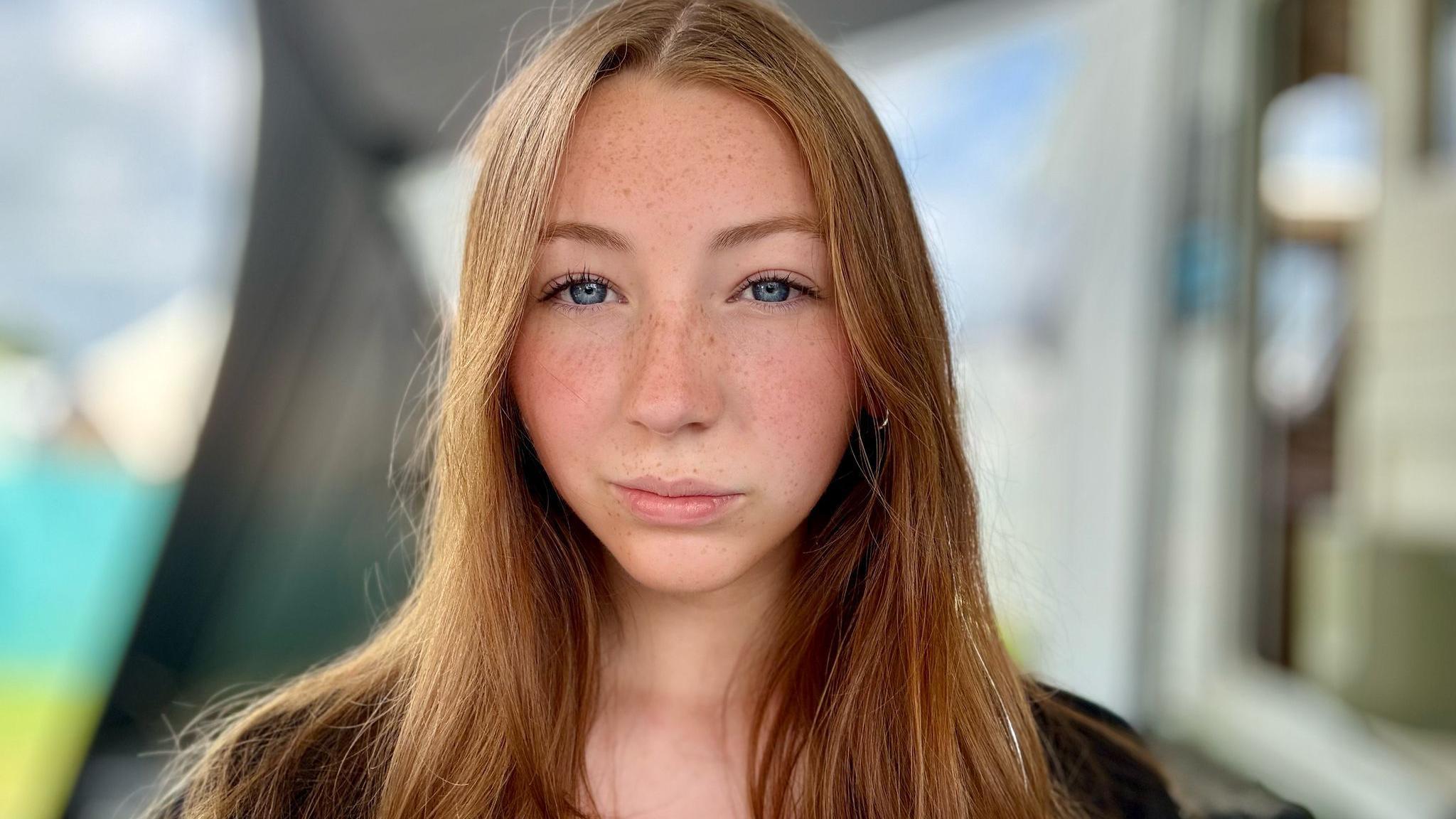 Abi has long brown hair and looks into the camera with a serious expression