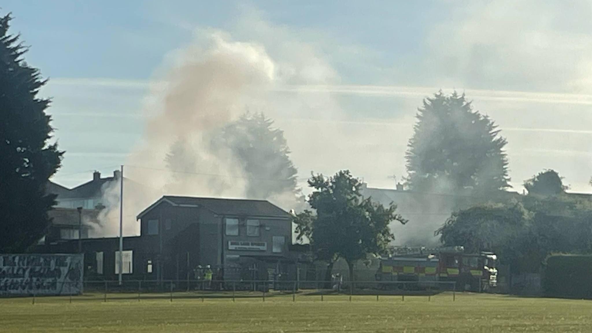 An image of a building in front of a large green area. Clouds of smoke are emerging behind the building and a fire engine is parked on the right next to a tree.