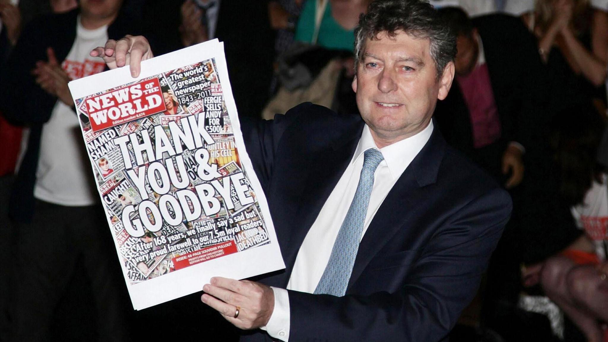 Colin Myler, the last editor of the News of the World, holds up its last edition as the paper was closed down in 2011. The paper front page says: Thank You & Goodbye. He is wearing a dark suit jacket, and a white shirt with a pale blue tie.