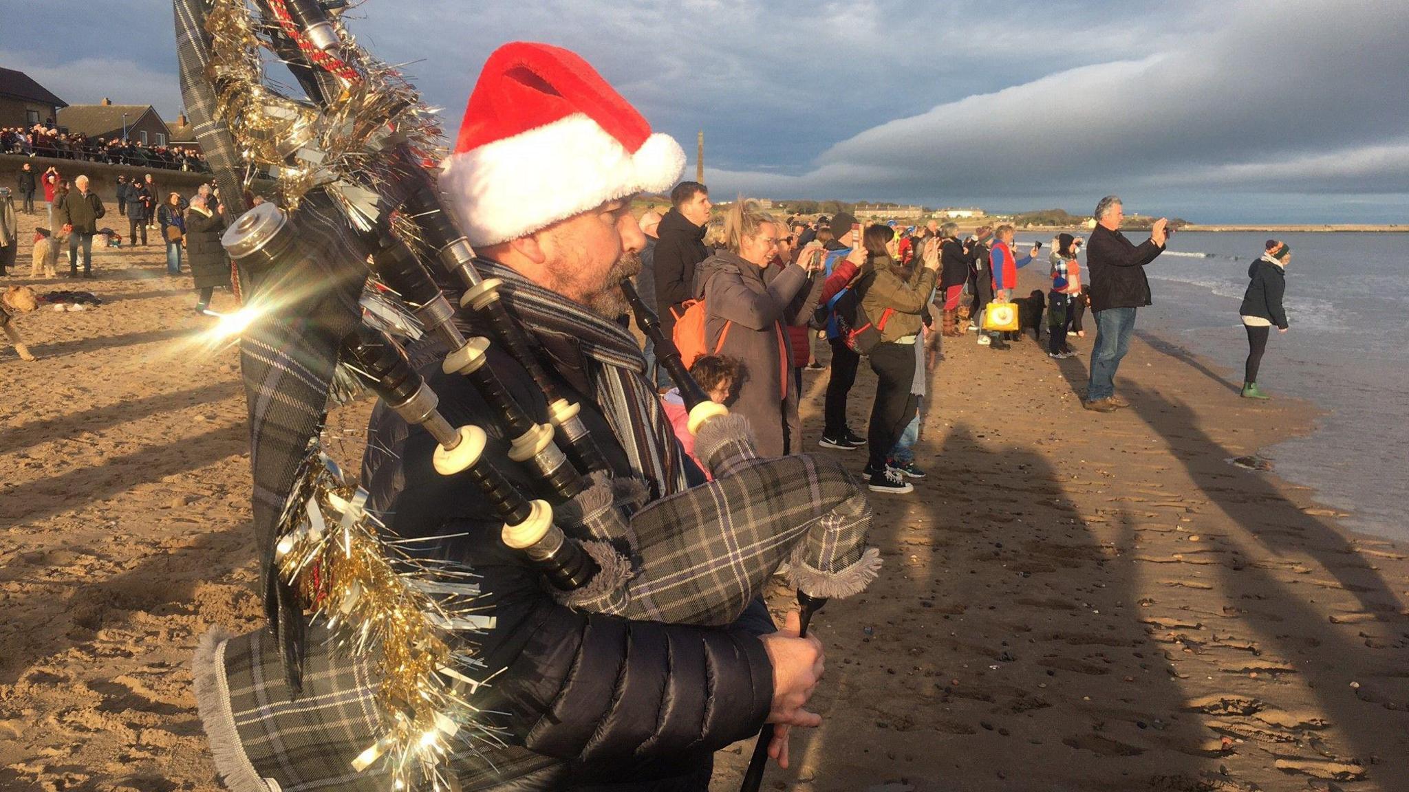 Piper Andrew Smith is standing on the sand at the water's edge playing his bagpipes. He is wearing tartan and a santa hat. There are dozens of people behind him taking snaps of the dippers in the water. 