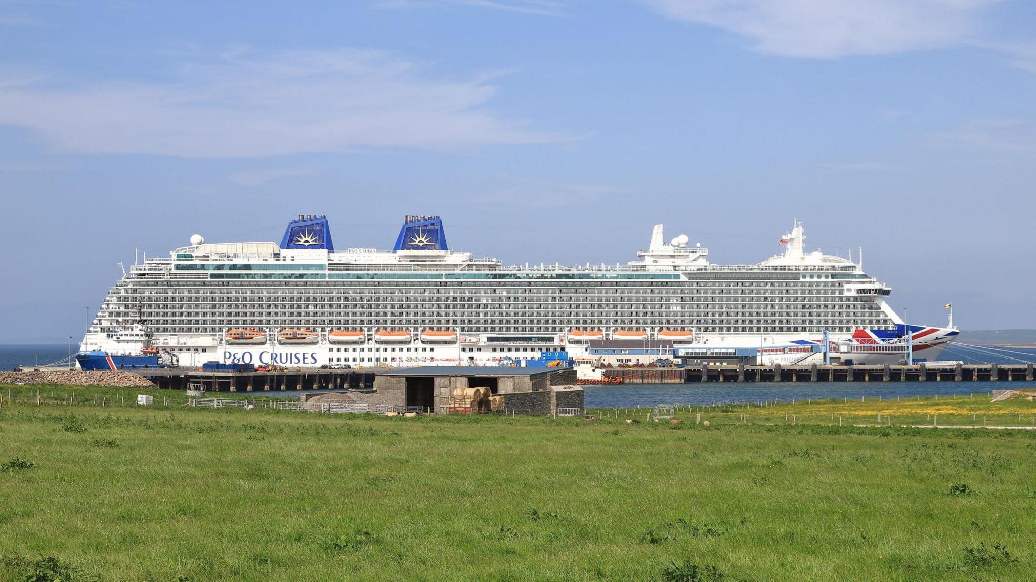 A large P&O cruise ship, Britannia, moored in Kirkwall in June 2023. A field of grass can be seen in the foreground.