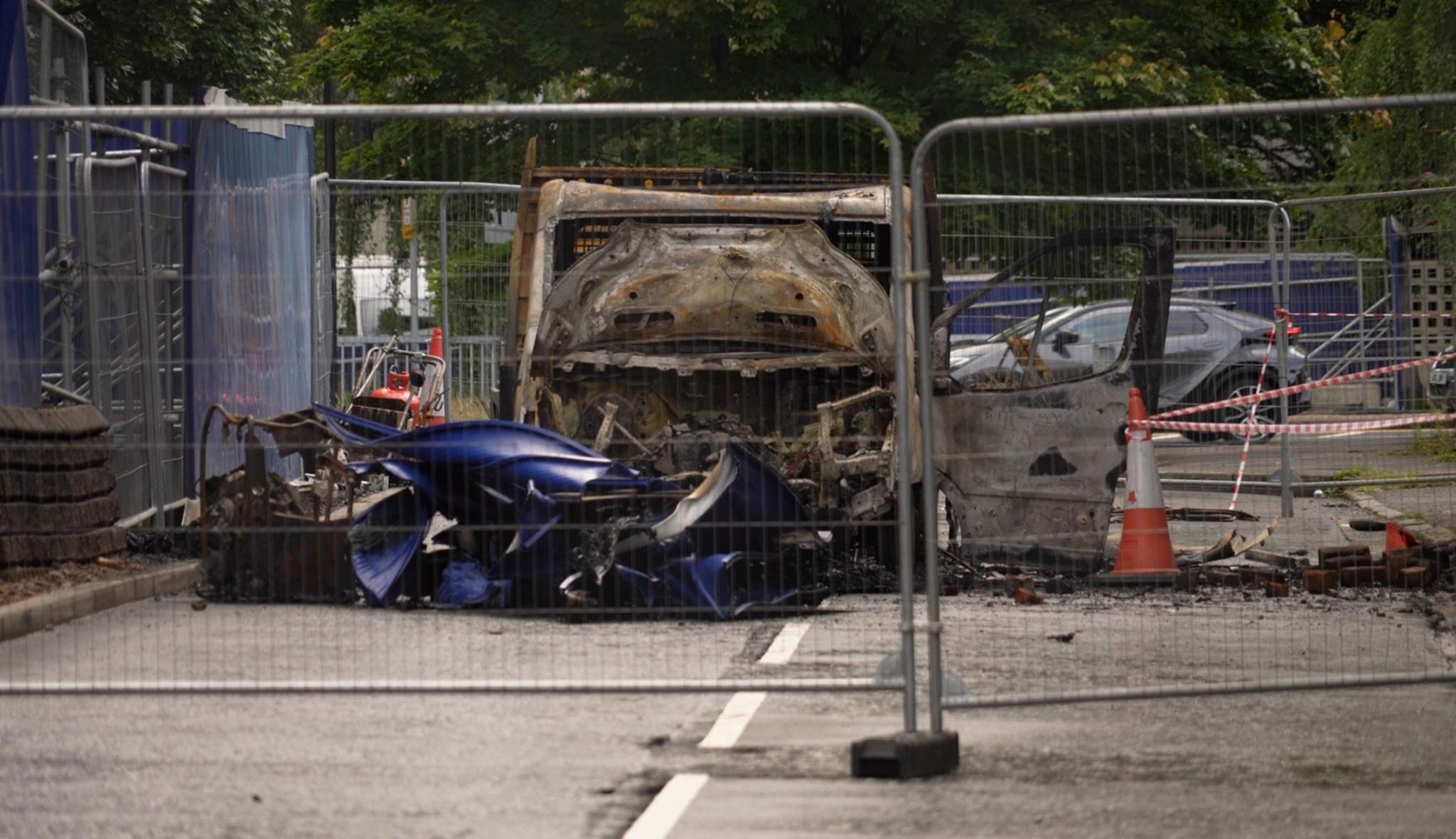 A destroyed truck, wrecked by fire, sitting on a city street. Fencing has been placed around the vehicle, and part of the road is dug up with traffic cones visible.