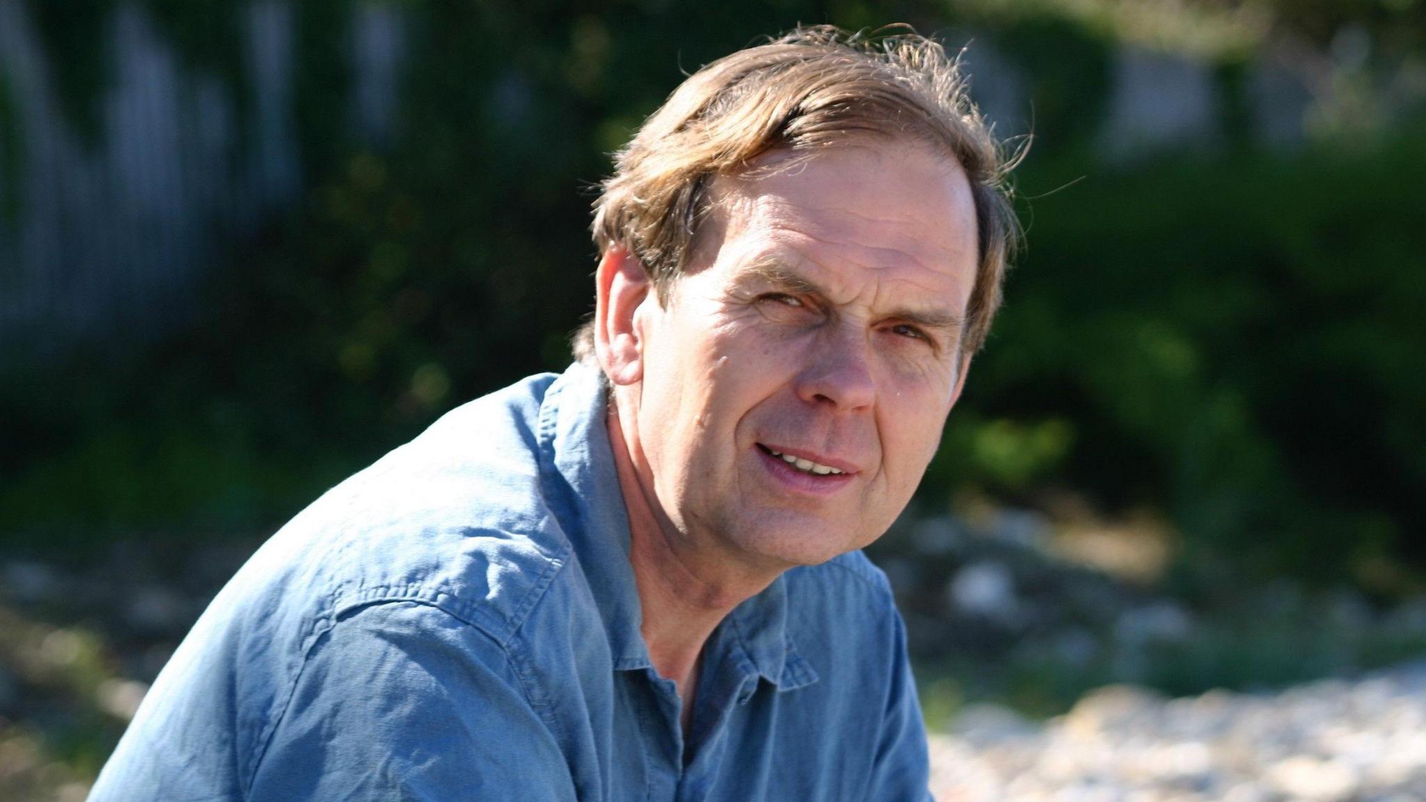 A man with light brown hair and wearing a blue shirt, looking slightly past the camera. It's a sunny day and he's squinting slightly.