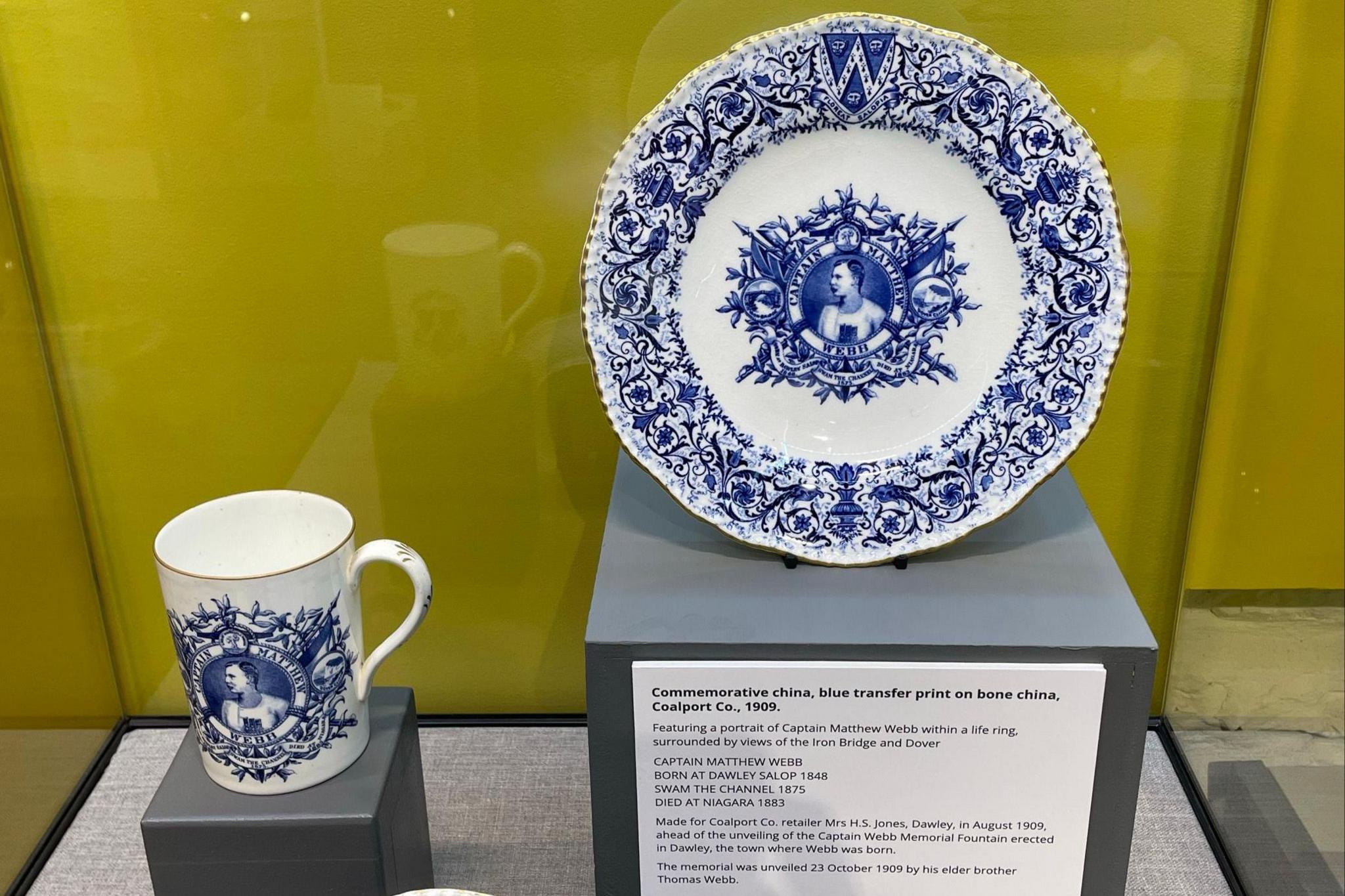 In a display case a white plate and cup both have blue floral designs around the edges with a motif of Captain Matthew Webb in the middle.