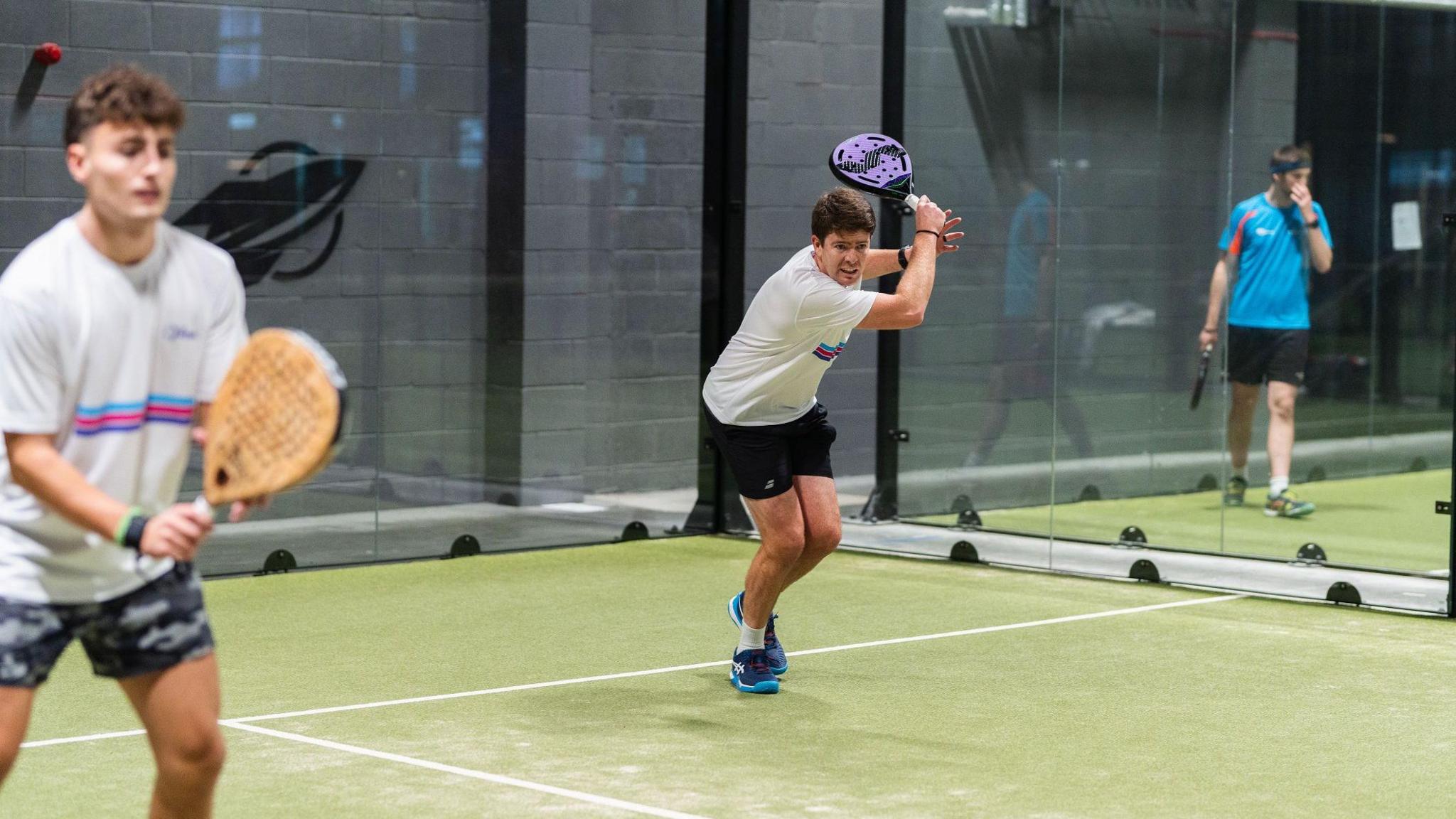 Three men holding sports rackets in a padel court. The playing surface is green with white lines