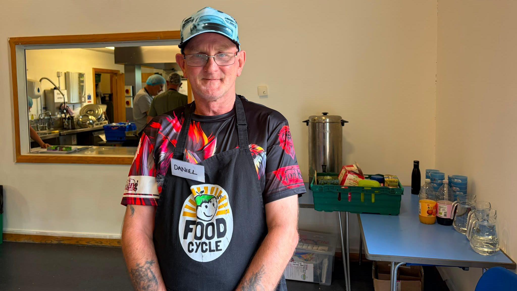 A tattooed man wearing a t-shirt with a red lion on an apron which says 'FoodCycle' is looking at the camera and smiling. Behind him is a table with water and squash on and a kitchen with people wearing hair nets.
