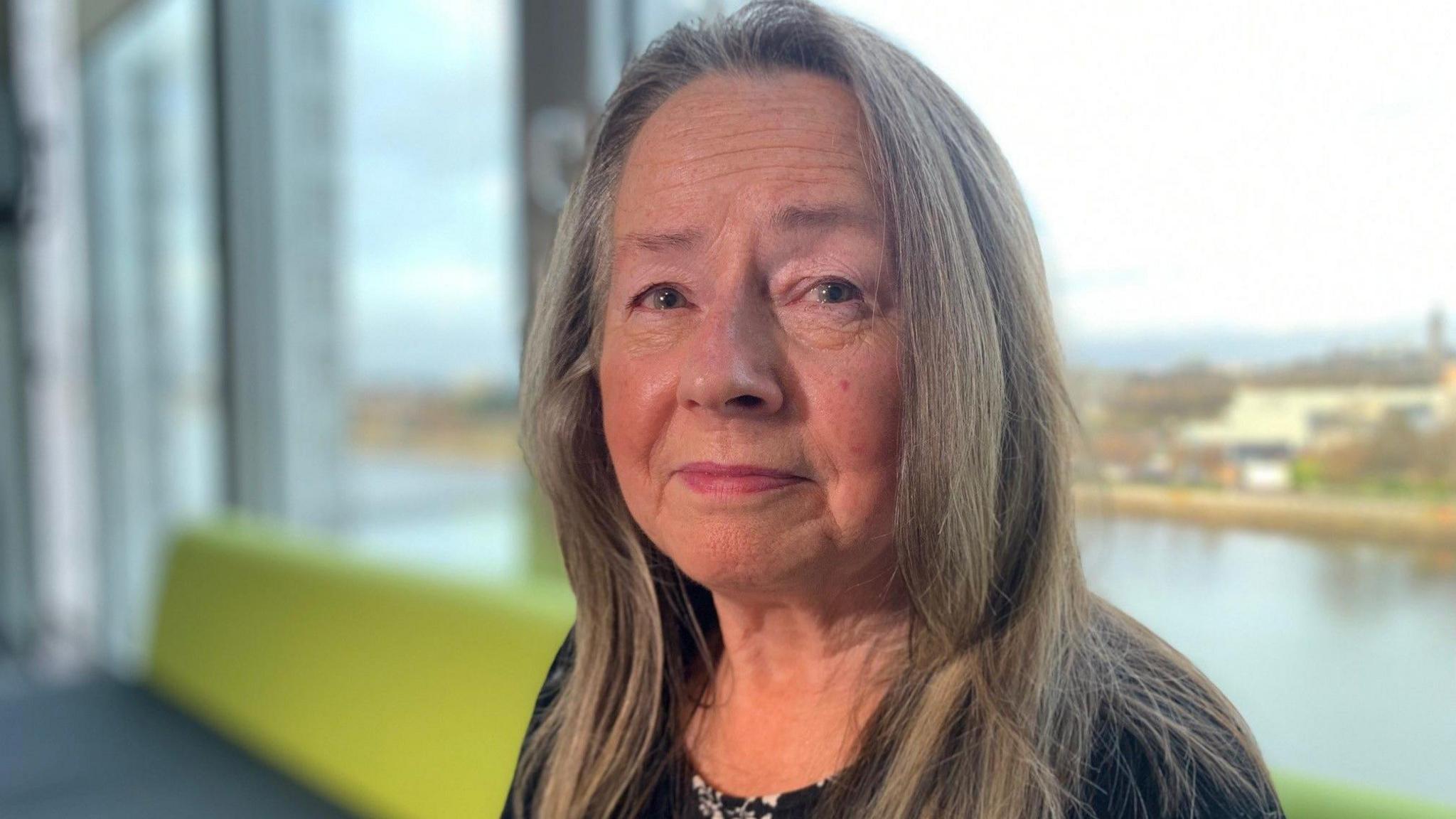 Image of Gina McGavin, a middle-aged woman with long light brown hair. She is sitting on a green sofa with a window running behind her - and a view of the River Clyde.