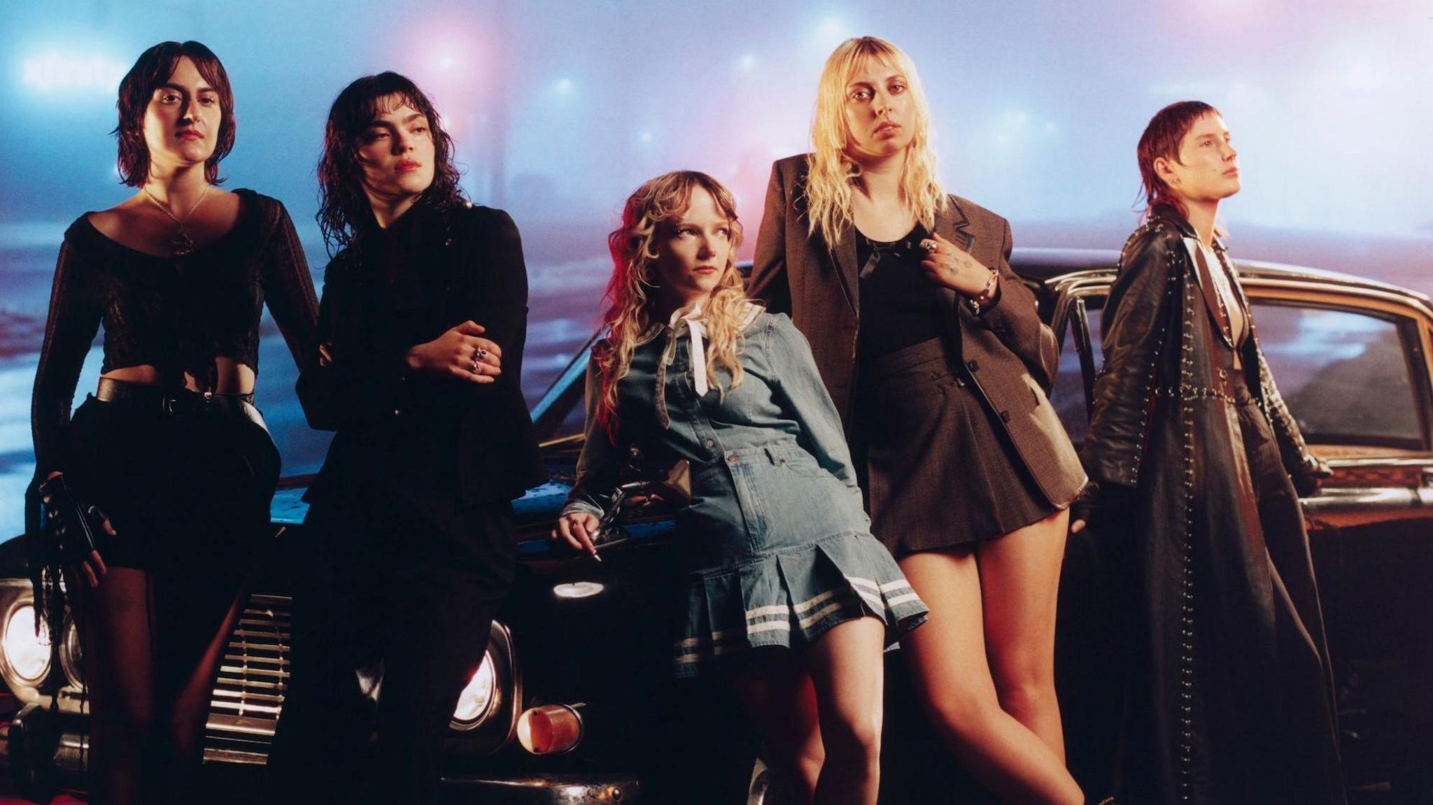 A promotional photo for The Last Dinner party shows the band leaning against a vintage car in a foggy urban landscape, illuminated by streetlamps