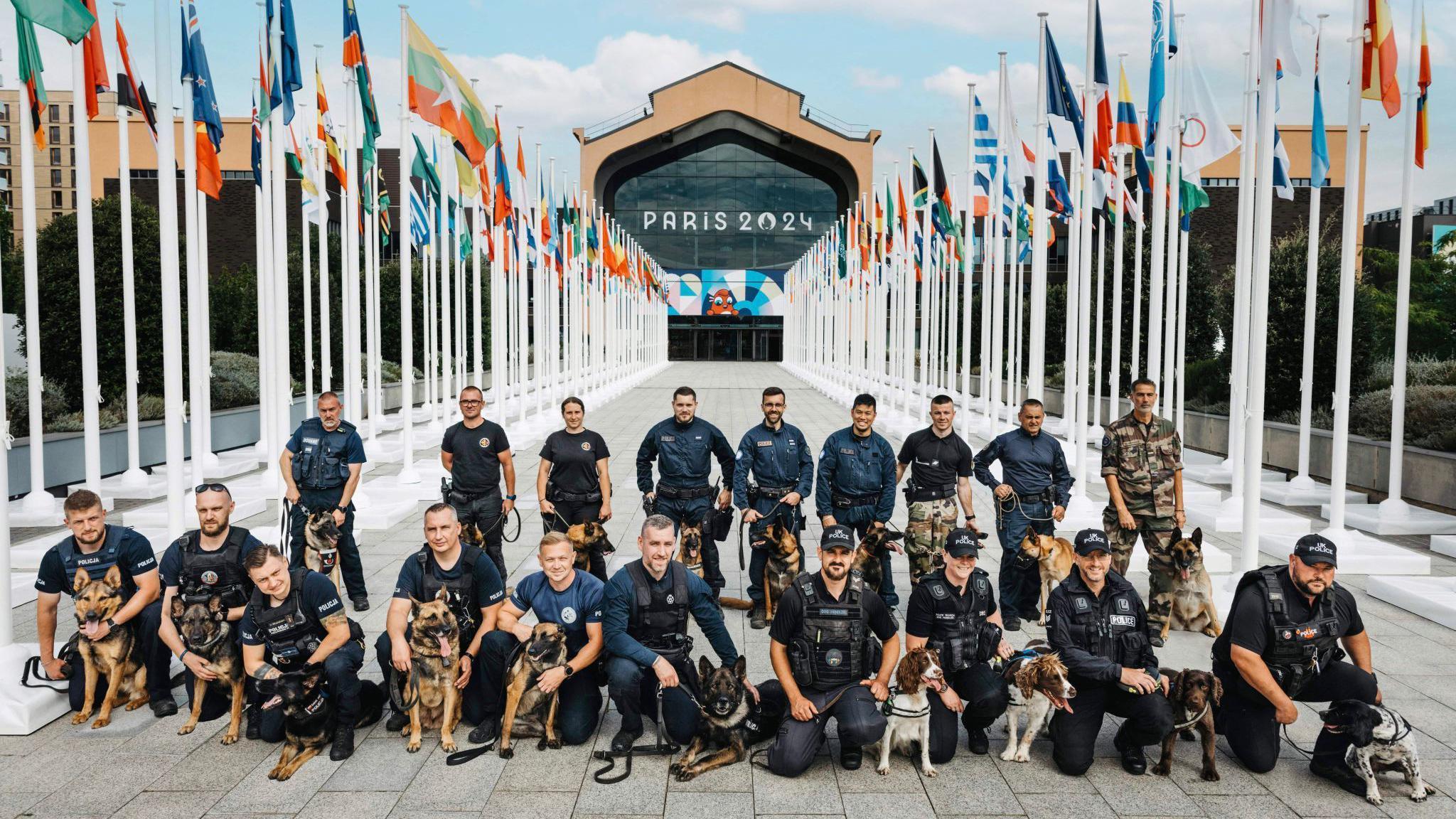 A group shot of policeman and their dogs kneeling in front of a building that says Paris 2024. They are surrounded by country's flags