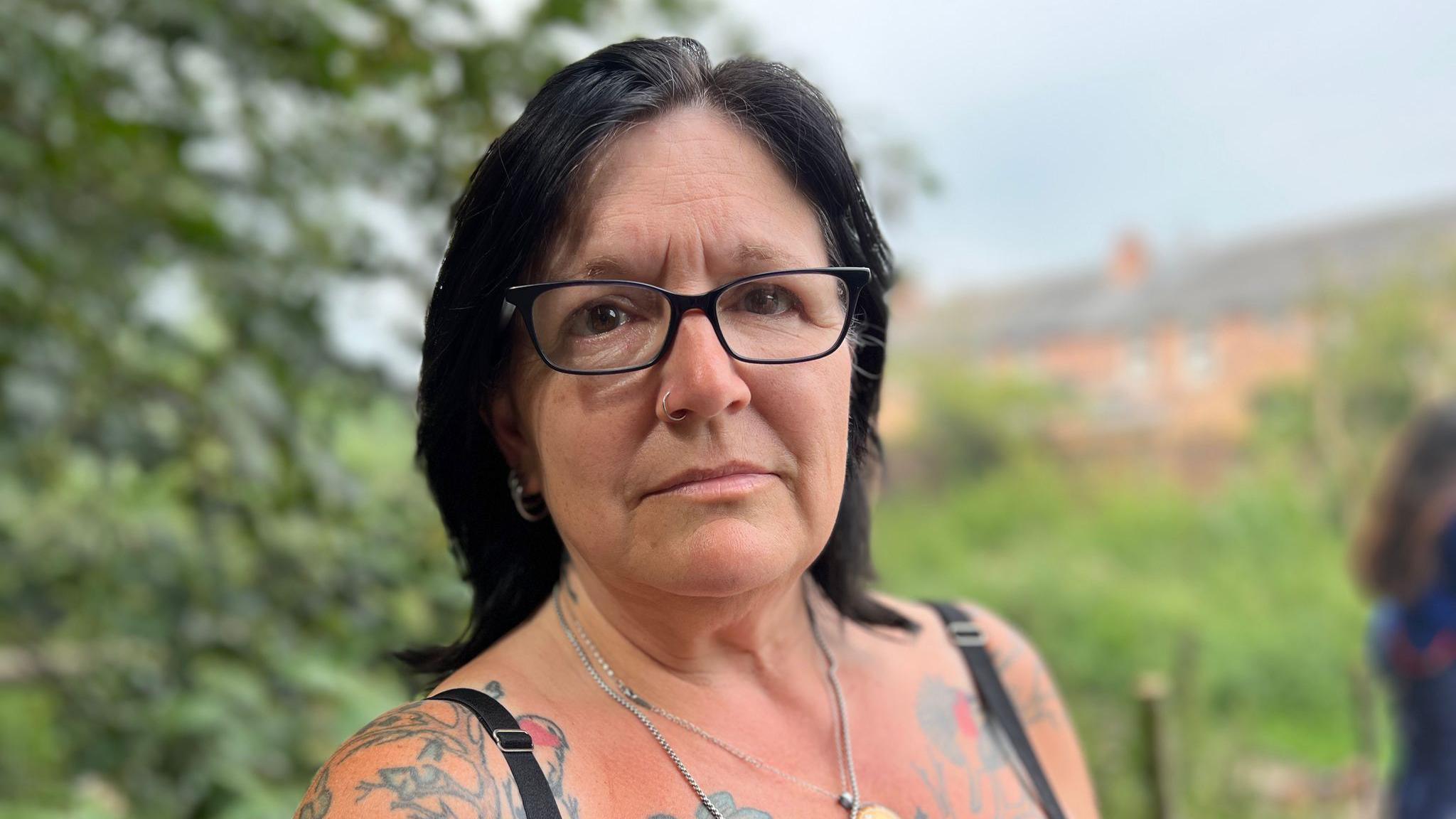 A woman with shoulder-length black hair and black rimmed glasses. She has a black vest on and has colourful tattoos on her arms and chest. She is stood in a field - green trees and grass can be seen blurred in the background.