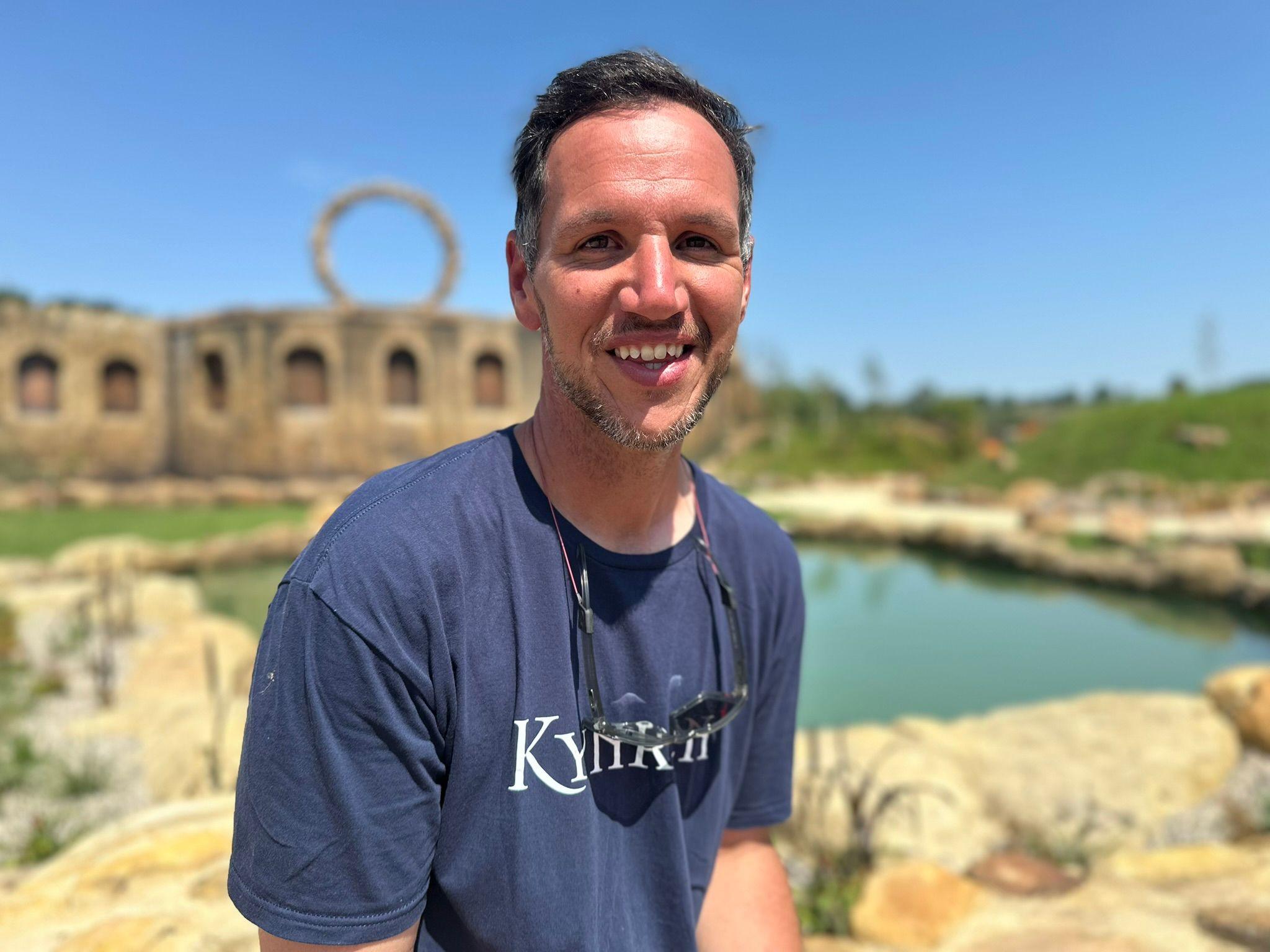 Jimmy Robinson smiles at the camera. He is wearing a navy tshirt with the word Kynren written across the front. He has short grey stubble and short dark hair and behind him is a large pond and stones.