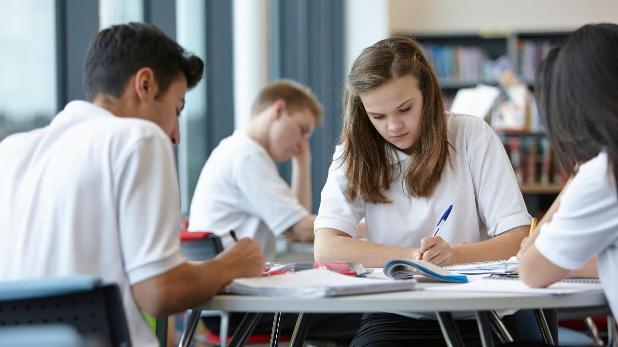 A group of students wearing white school uniforms sit at a table in a classroom, working on assignments. Open notebooks and pens are visible on the table. Bookshelves filled with books line the background, and large windows allow in natural light.