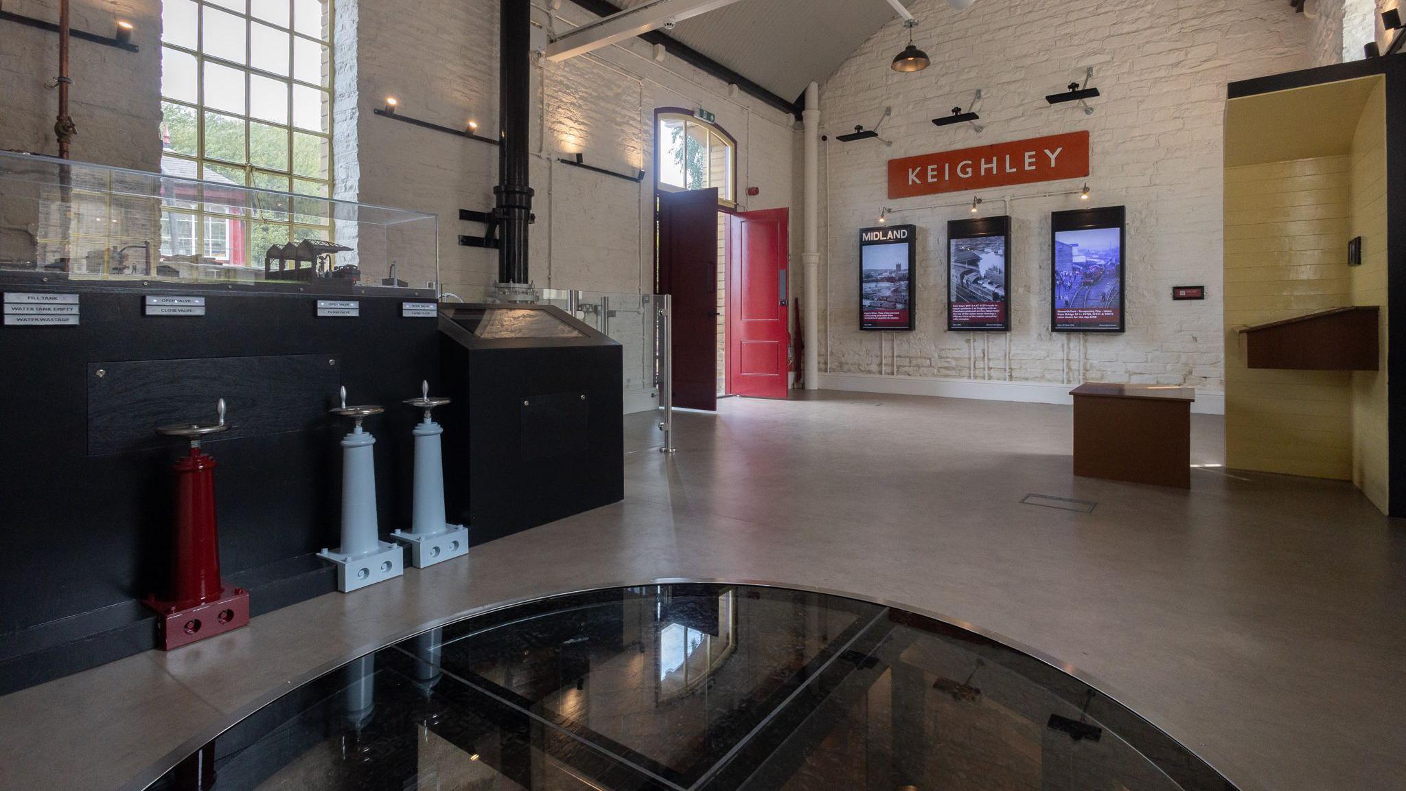The interior of the visitor centre. The walls are painted white and constructed from stone or brick. The ceiling is arched and features exposed beams painted in a dark colour, along with modern lighting fixtures hanging down.
On the left side of the image, there is a large window with multiple panes. Below the window, there are display cases containing various objects, including three tall, narrow structures in red, grey, and blue, which appear to be historical railway equipment or signalling devices. Each display has labels for identification.
In the centre foreground, there is a circular glass-covered feature set into the floor. Toward the back wall, there is a bright red door that stands open. Above this door, there is a sign that reads “KEIGHLEY”. Below the sign, three digital display screens are mounted on the wall, showing images and text related to railway history.
