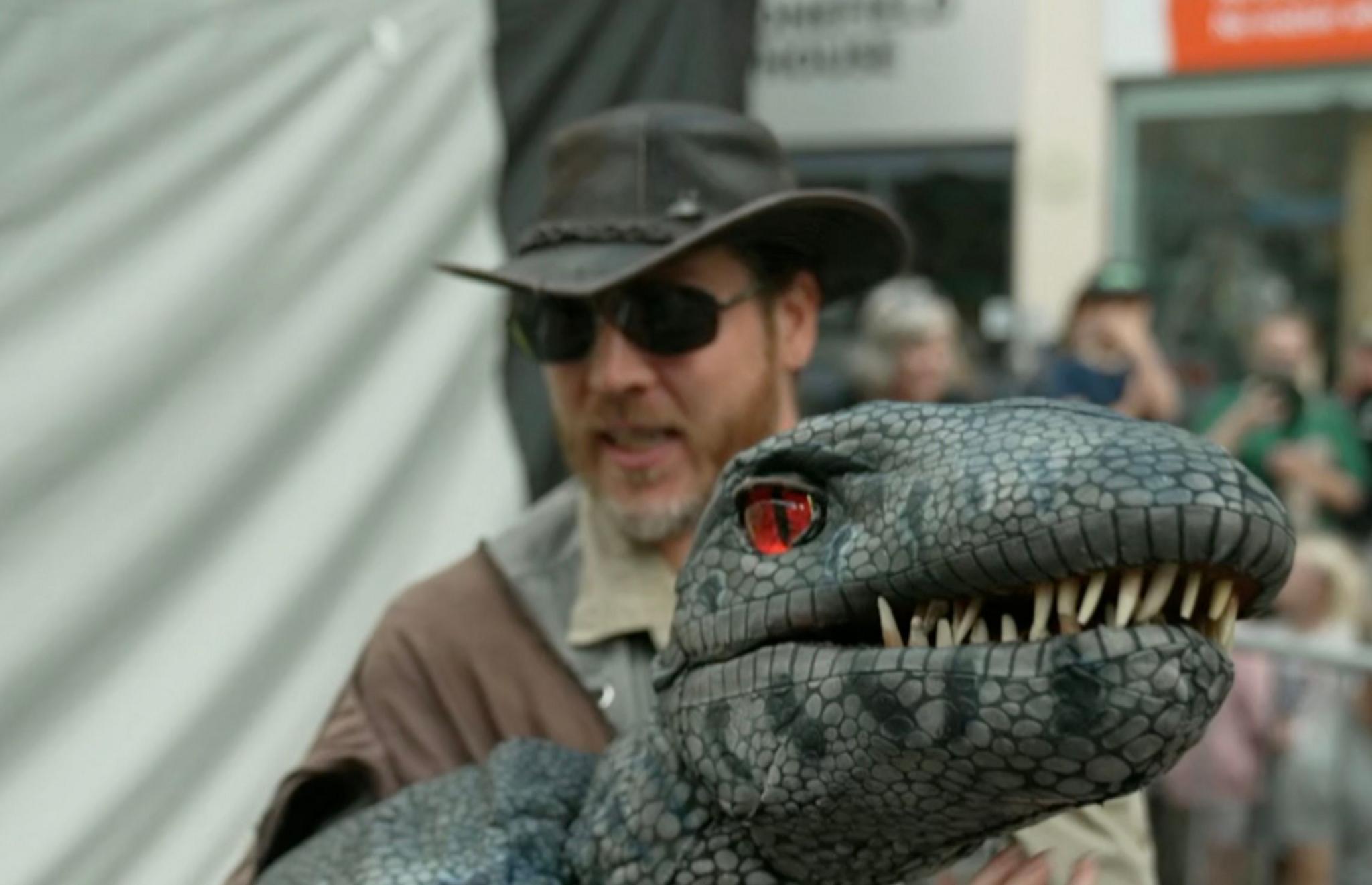 A grey-blue dinosaur puppet with a red eye and white teeth. In the background, out of focus, is a man wearing a brown safari hat, sunglasses and safari-style clothes.