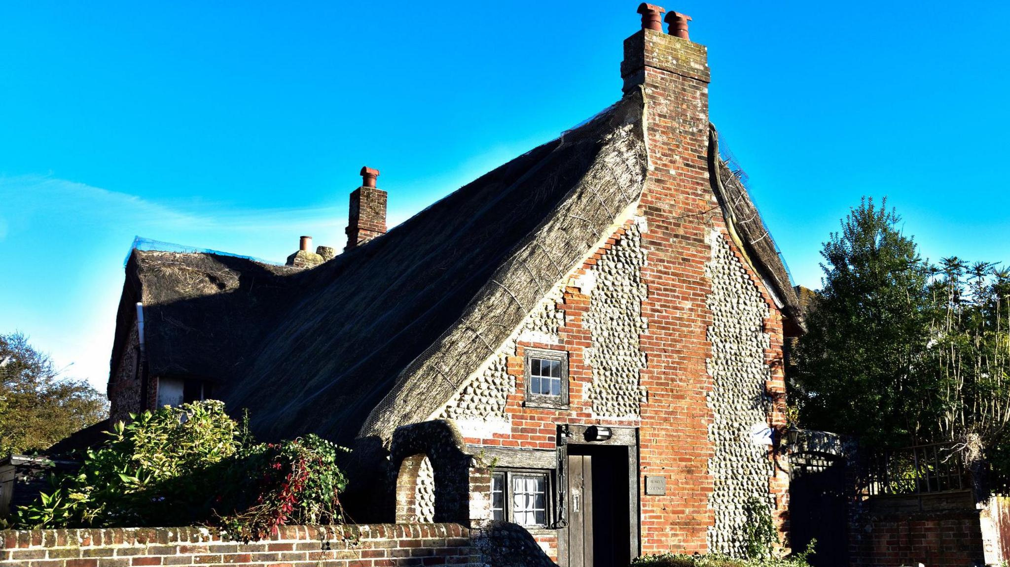 A thatched roof building with red brick and grey stones. The cottage has two small windows and a wooden door. 