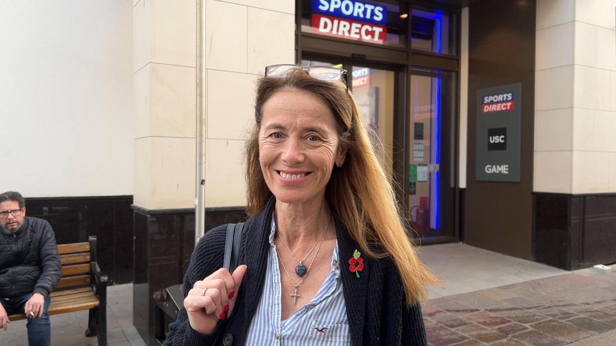 Gayla Bridge, a woman with shoulder-length light-brown hair. She has a pair of glasses on her head. She is wearing a black pullover with a poppy on the lapel, over a blue and white striped shirt. She is also wearing two necklaces, and has a bag over one of her shoulders. Behind her is the entrance to a sports shop. To the left of the image, a man is visible sitting on a bench.