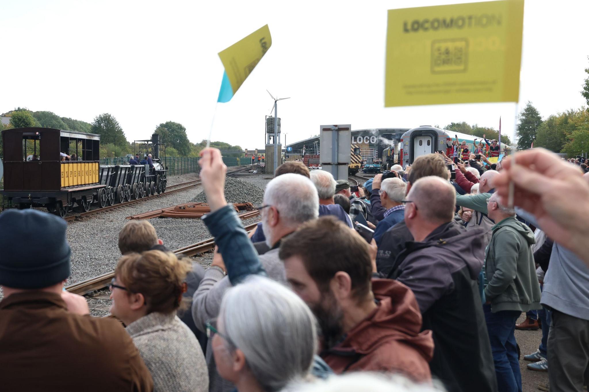 A large crowd of people cheers and waves yellow and blue flags as a black steam engine 