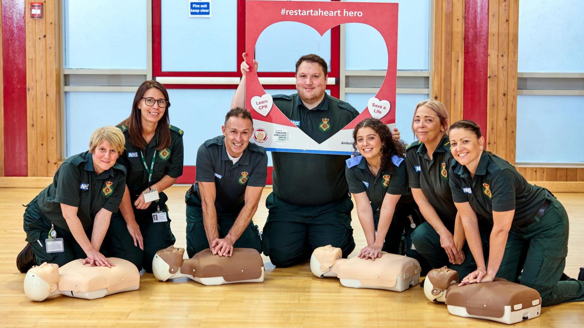 Seven uniformed ambulance workers kneel on a gymnasium floor, actively engaged in CPR training. Each person practises chest compressions on a dummy, smiling and focused. One participant holds a red heart-shaped frame with the hashtag “#restartaheart hero” and messages promoting CPR awareness, such as “Learn CPR” and “Save a Life".