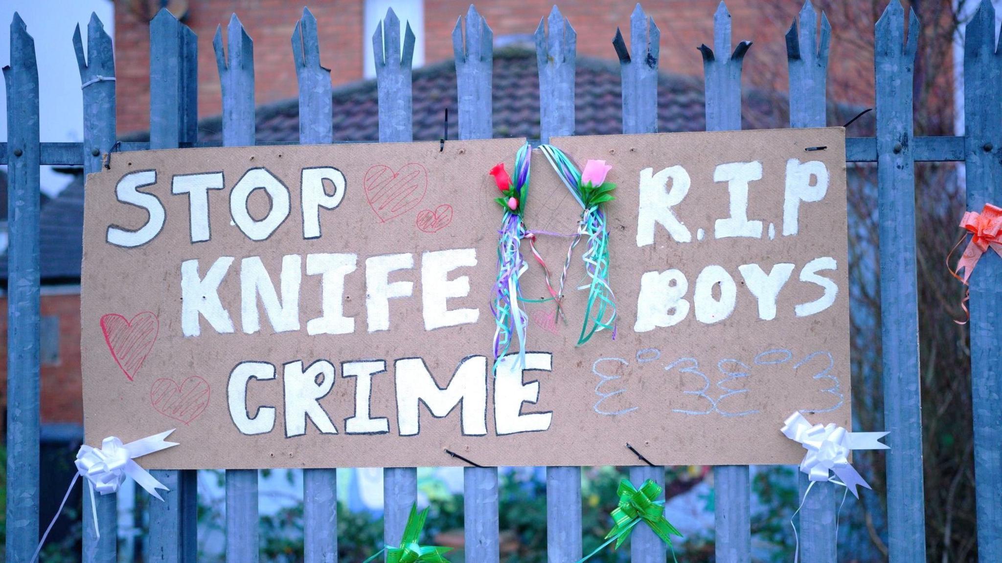 A handmade cardboard sign attached to a spiked metal fence on a pavement. It says "Stop Knife Crime. RIP Boys". Several coloured ribbons and flowers have also been tied to it. 